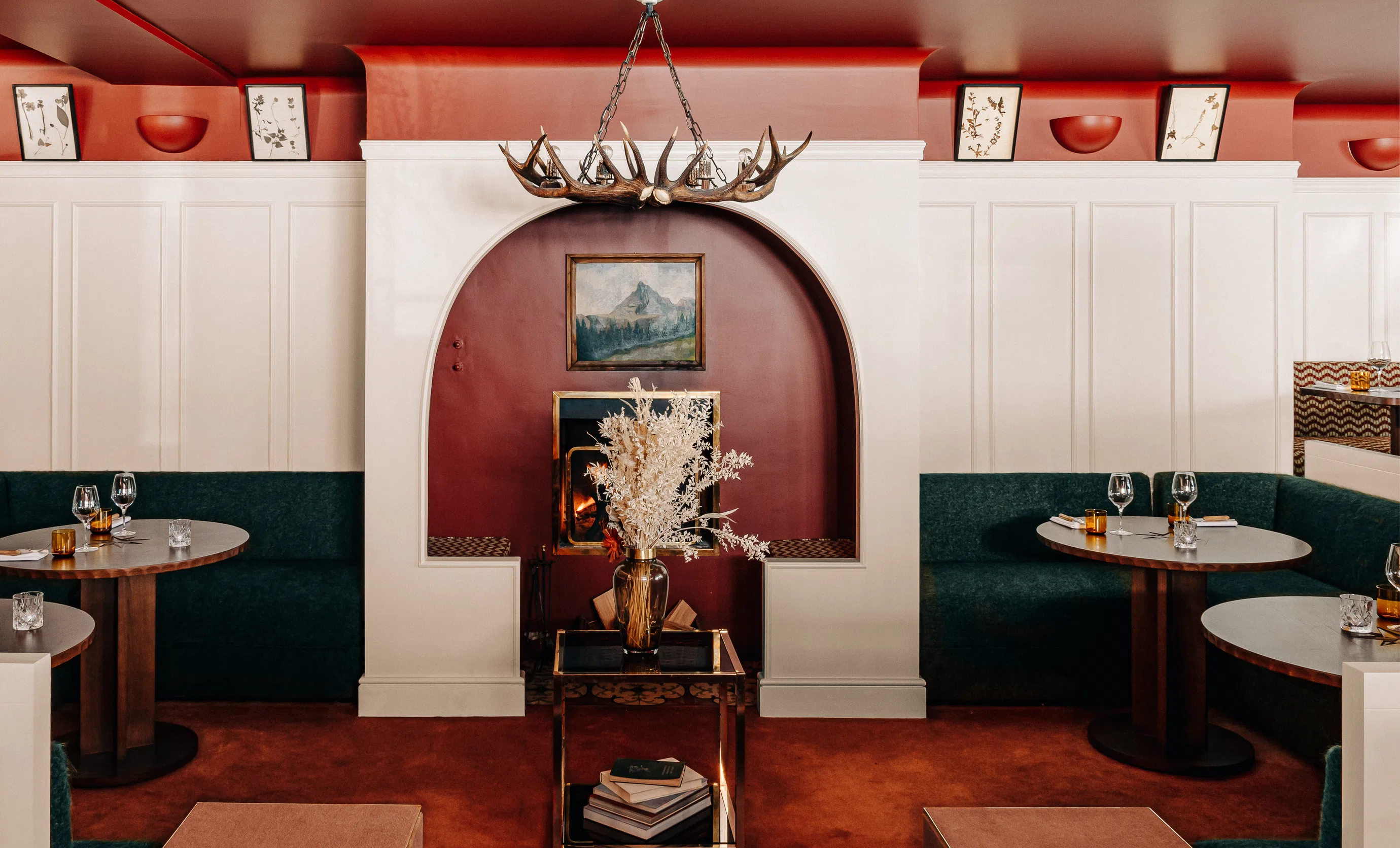 Cozy restaurant interior with dark green bench seating, round wooden tables set with glassware, a fireplace with antler chandelier, and a vase of dried flowers on a glass table.
