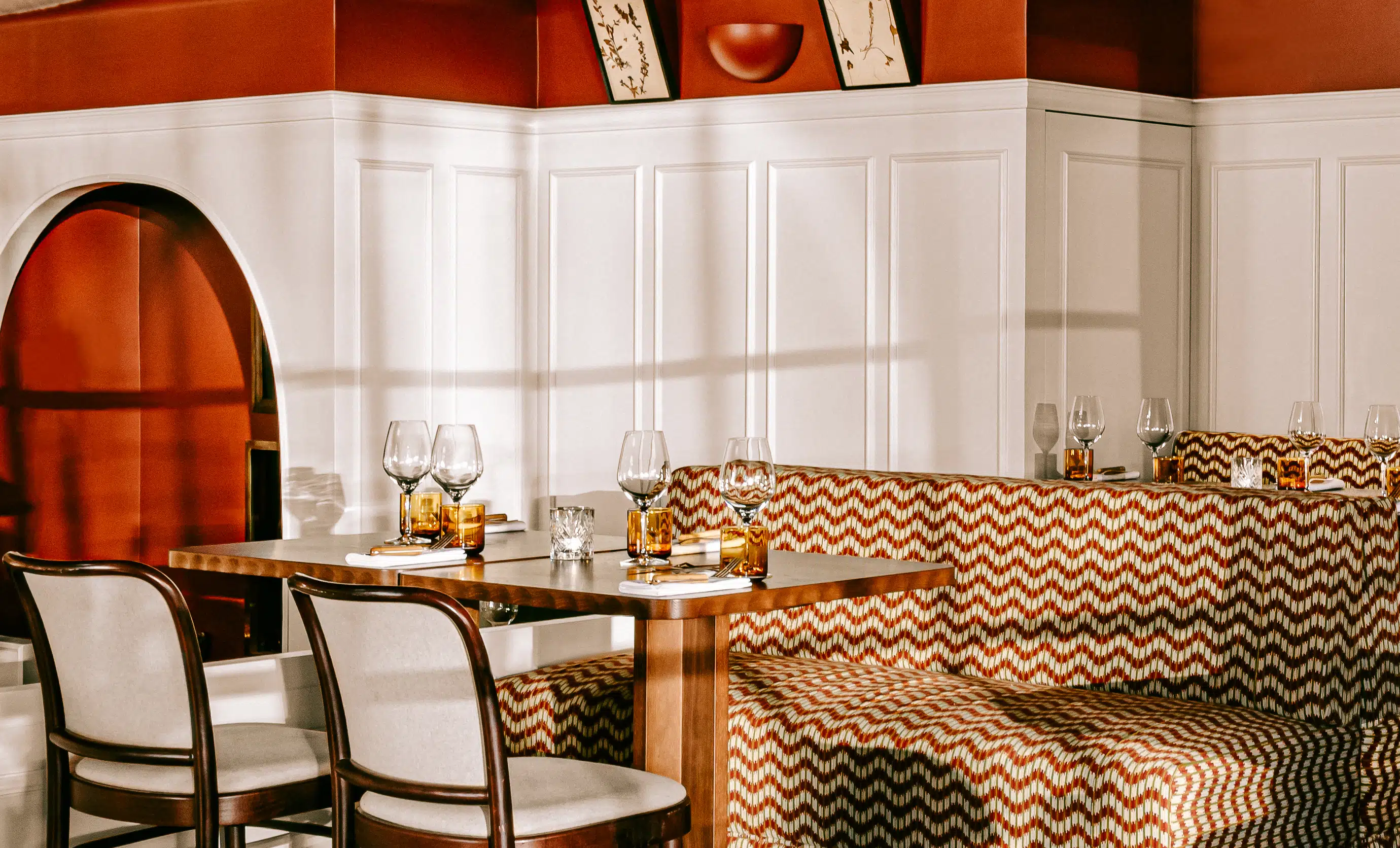 Elegant dining area with wooden table, patterned orange and white cushioned bench seating, beige chairs, and wine glasses on table.