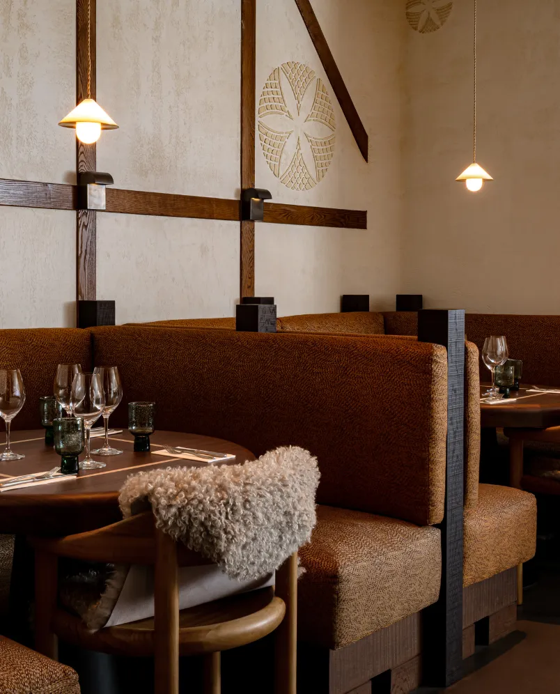 Cozy restaurant booth seating with brown upholstery, wooden chairs draped with a soft woolen throw, round tables set with wine glasses and utensils, under warm pendant lighting.