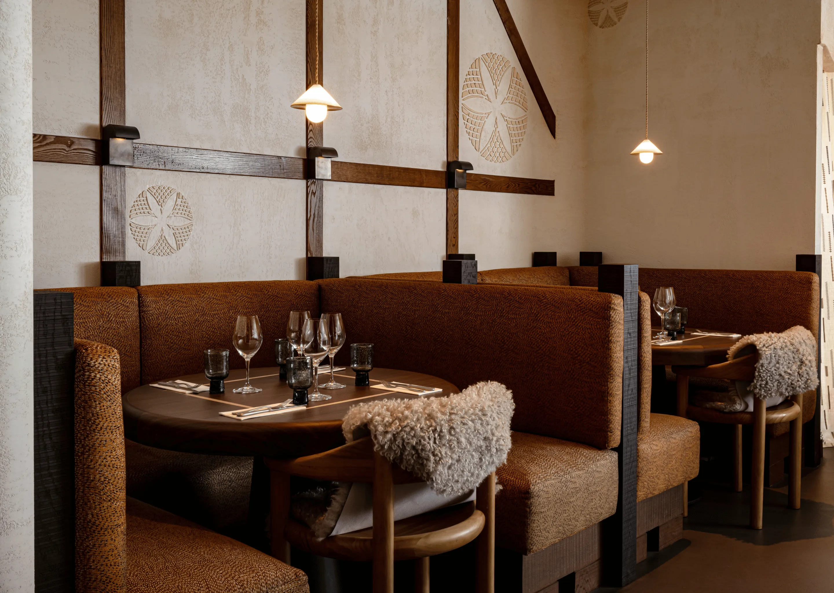 Cozy restaurant booths with brown upholstery, wooden tables set with wine glasses, and wooden chairs draped with beige sheepskin throws under hanging pendant lights.