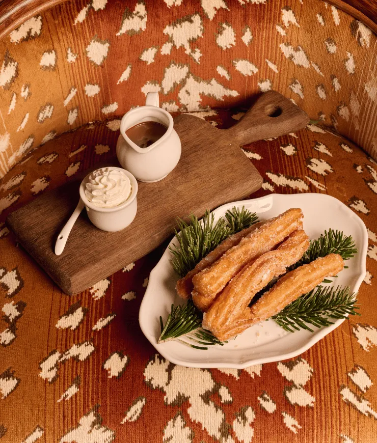 Plate of sugar-coated churros garnished with pine sprigs, served with a white cup of whipped cream and a small pitcher of chocolate sauce on a wooden board.