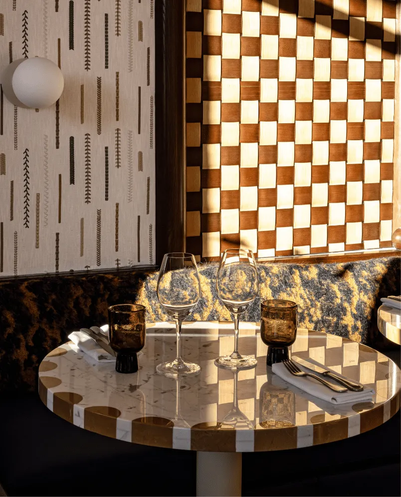 Round marble table with gold and white checkered edges set with two wine glasses, two brown water glasses, and cutlery on white napkins in a sunlit restaurant corner with patterned walls and cushioned bench.