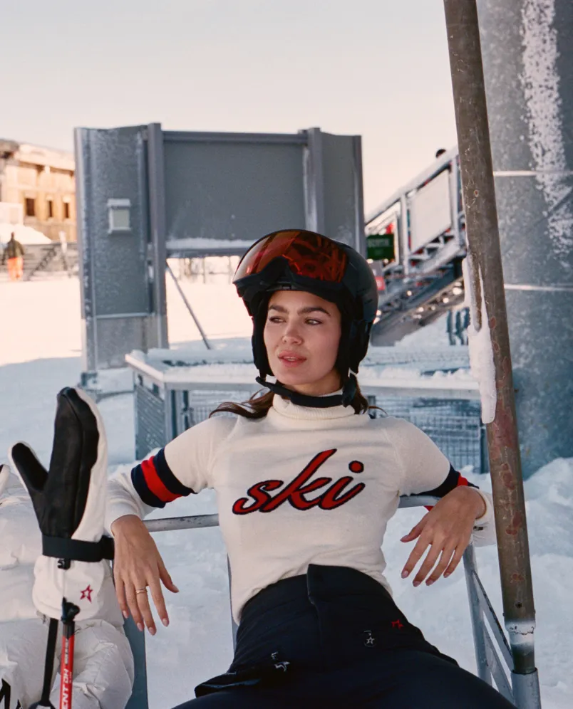 Woman wearing ski helmet and white sweater with 'ski' text, sitting on a ski lift at a snowy resort.