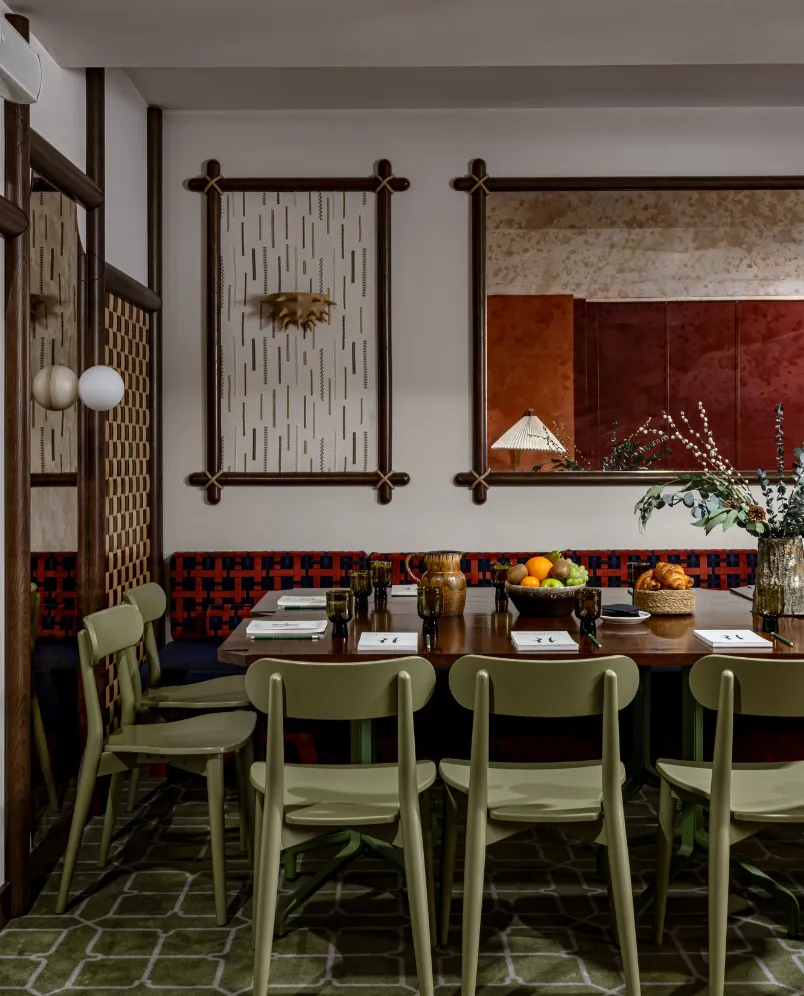 Dining room with olive green chairs around a wooden table set with drinks, notebooks, and fruit bowls under a large mirror and decorative wall art.