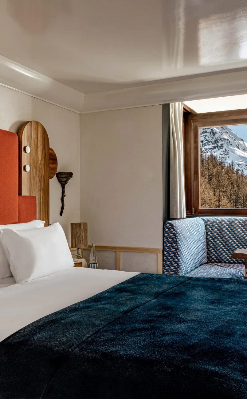 Hotel room with white pillows, an orange headboard, blue patterned seating by a window showing snowy mountain and trees.