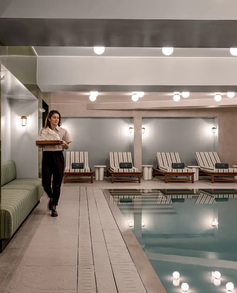 Woman carrying towels walking beside an indoor pool with striped lounge chairs and soft lighting.