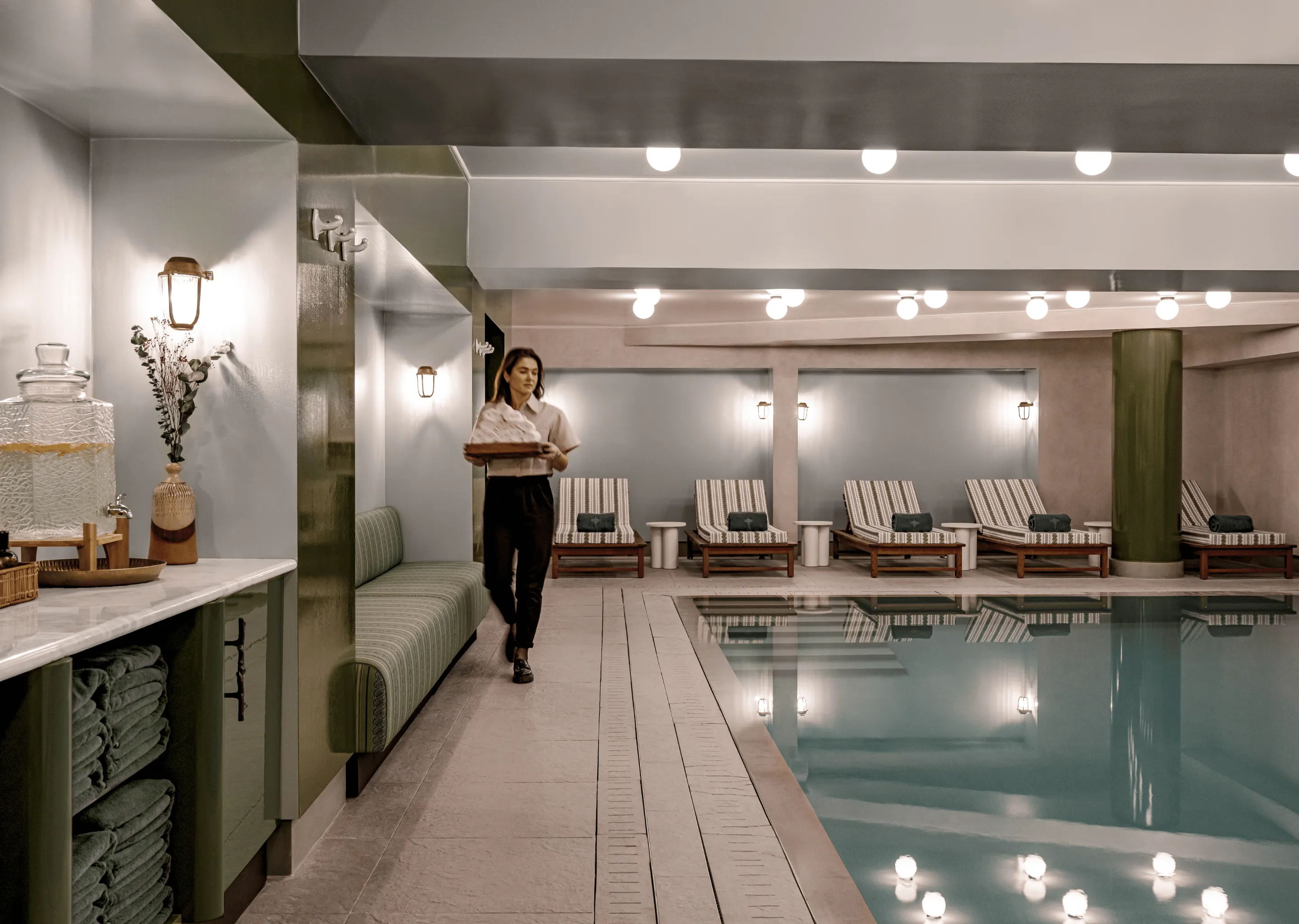 Indoor wellness pool area with striped lounge chairs, rolled towels, ambient lighting, and a person carrying towels on a tray.