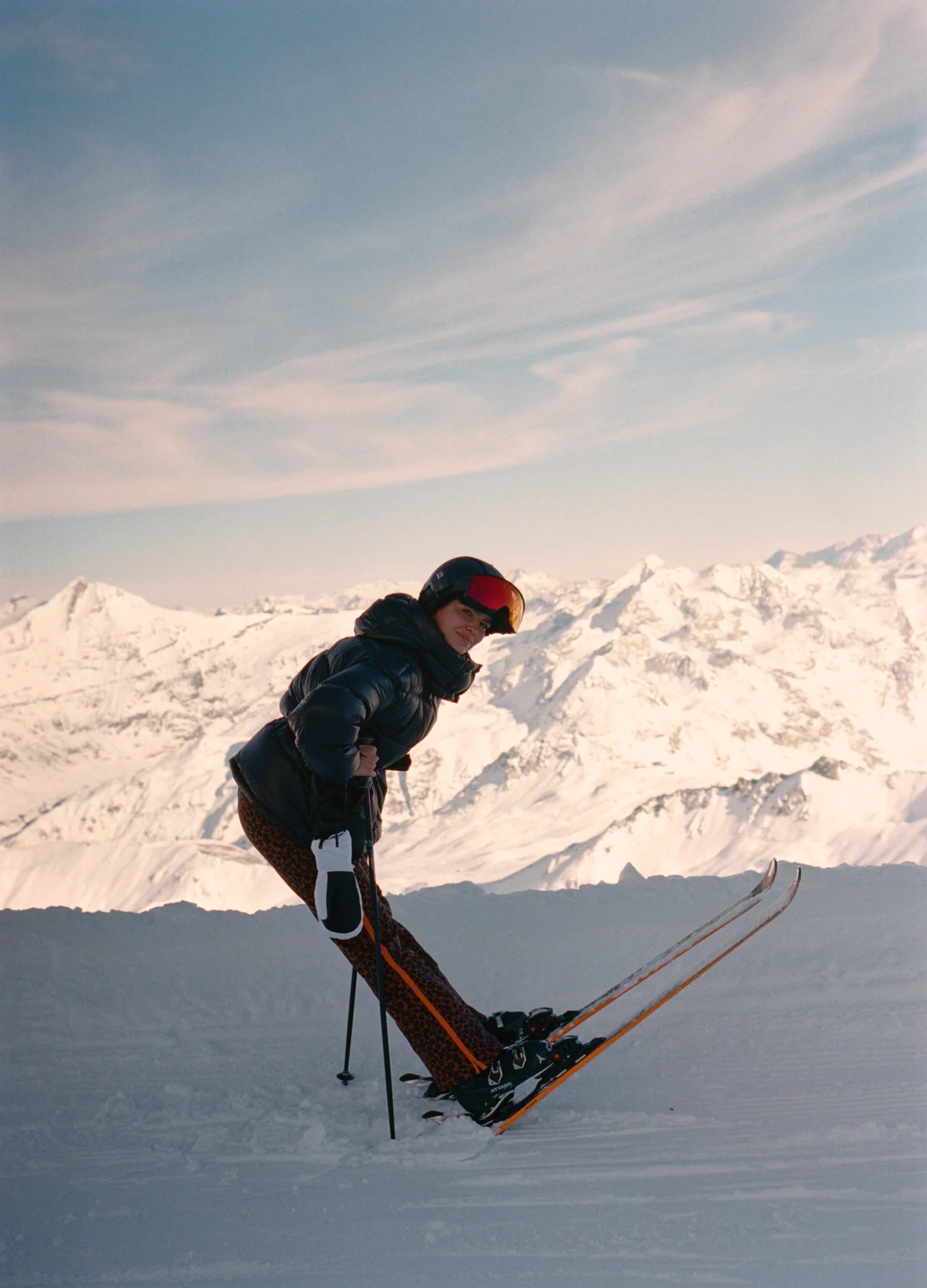 Skier wearing a black helmet and puffer jacket leaning forward on skis with snowy mountain backdrop under a blue sky.
