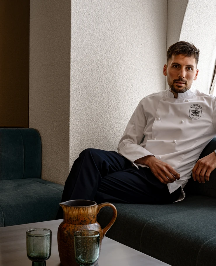 Man wearing a white chef jacket sitting on a dark green sofa next to a table with two green glasses and a brown ceramic pitcher.
