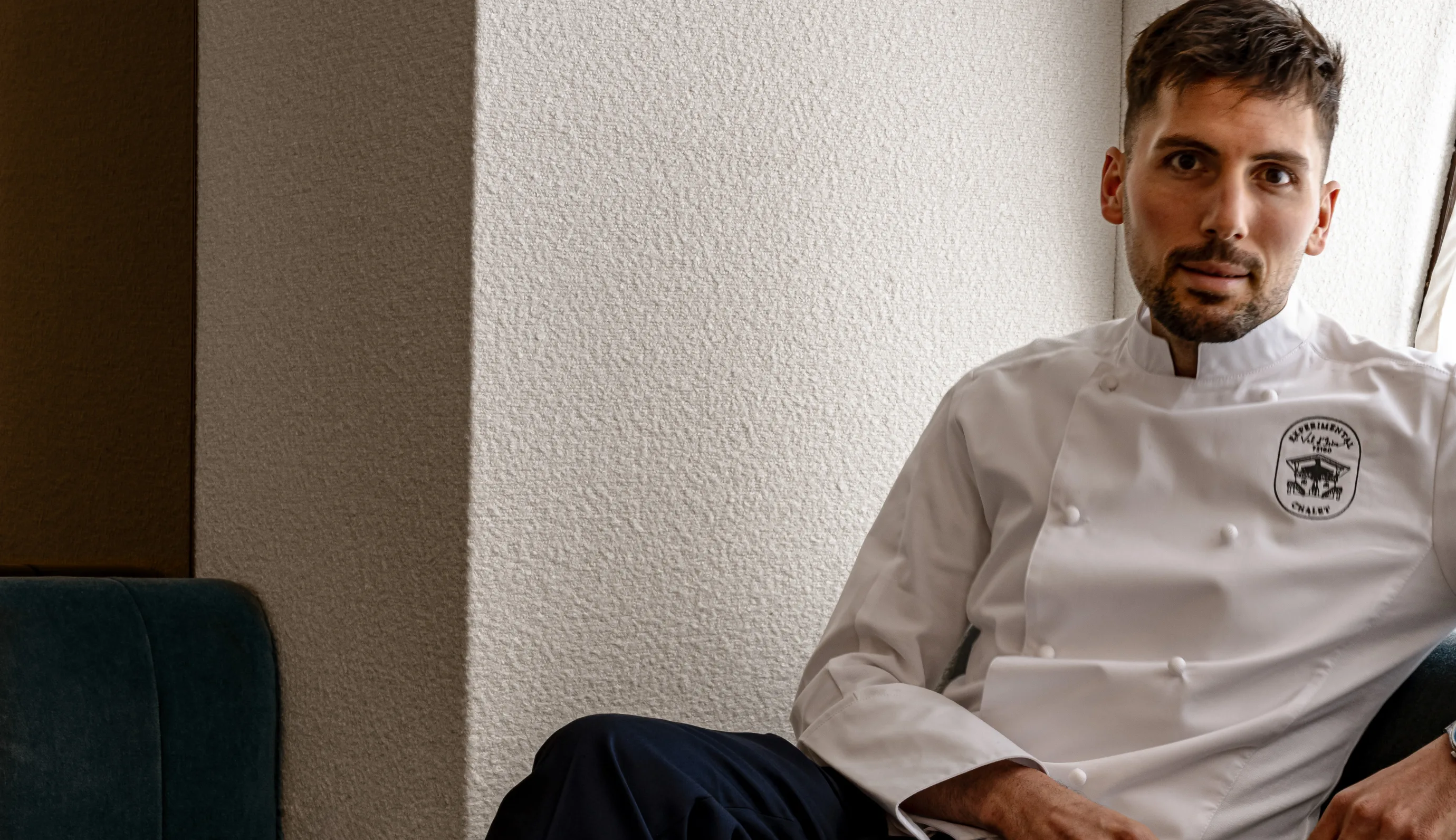Man with short brown hair and beard wearing a white chef jacket with an embroidered logo, sitting against a textured beige wall.
