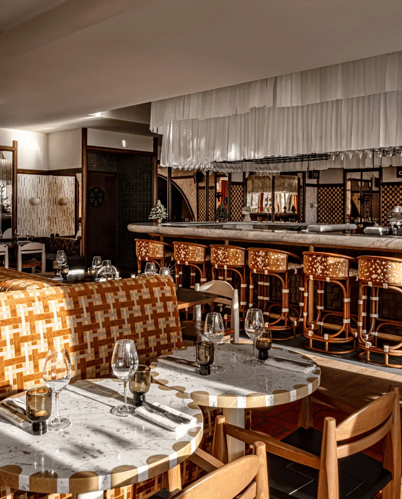 Cozy restaurant interior featuring marble tables set with glassware, sunlit patterned chairs, and a bar with floral-patterned bar stools under hanging glassware.
