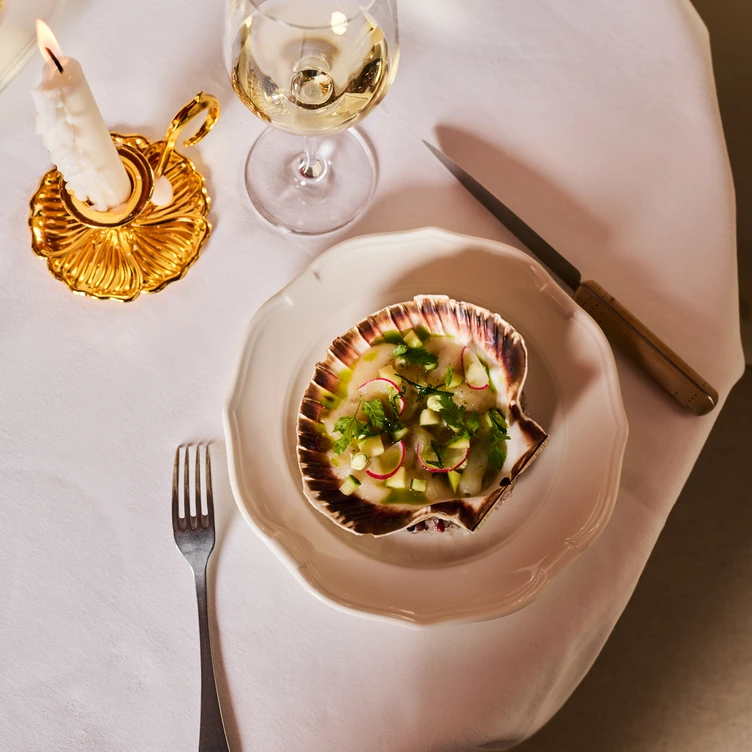 Gourmet appetizer served in a scallop shell on a plate, next to a fork, a knife, a candle in a gold holder, and a glass of white wine on a white tablecloth.