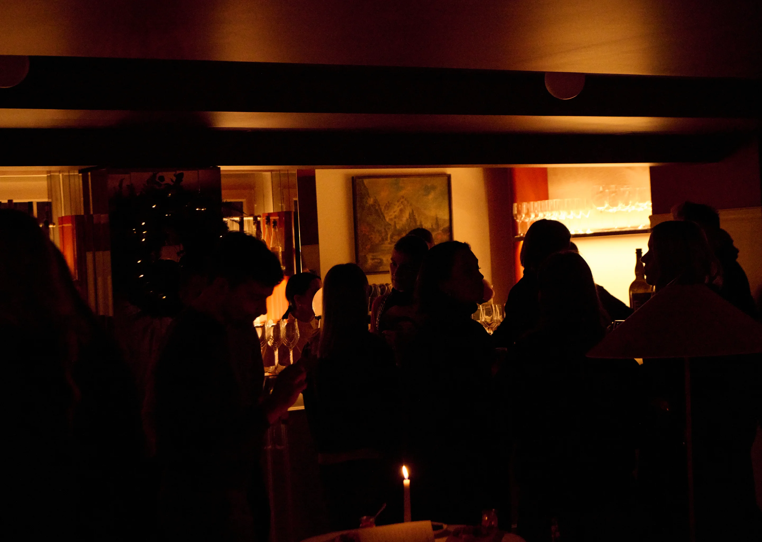 Silhouettes of people socializing in a dimly lit room with a lit candle on a table and shelves of wine glasses in the background.