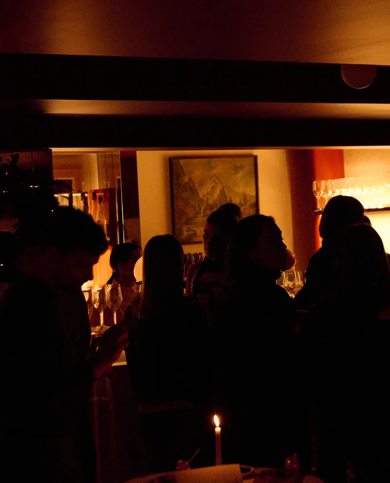 Dimly lit room with a group of people socializing near a candle on a table and shelves with wine glasses.