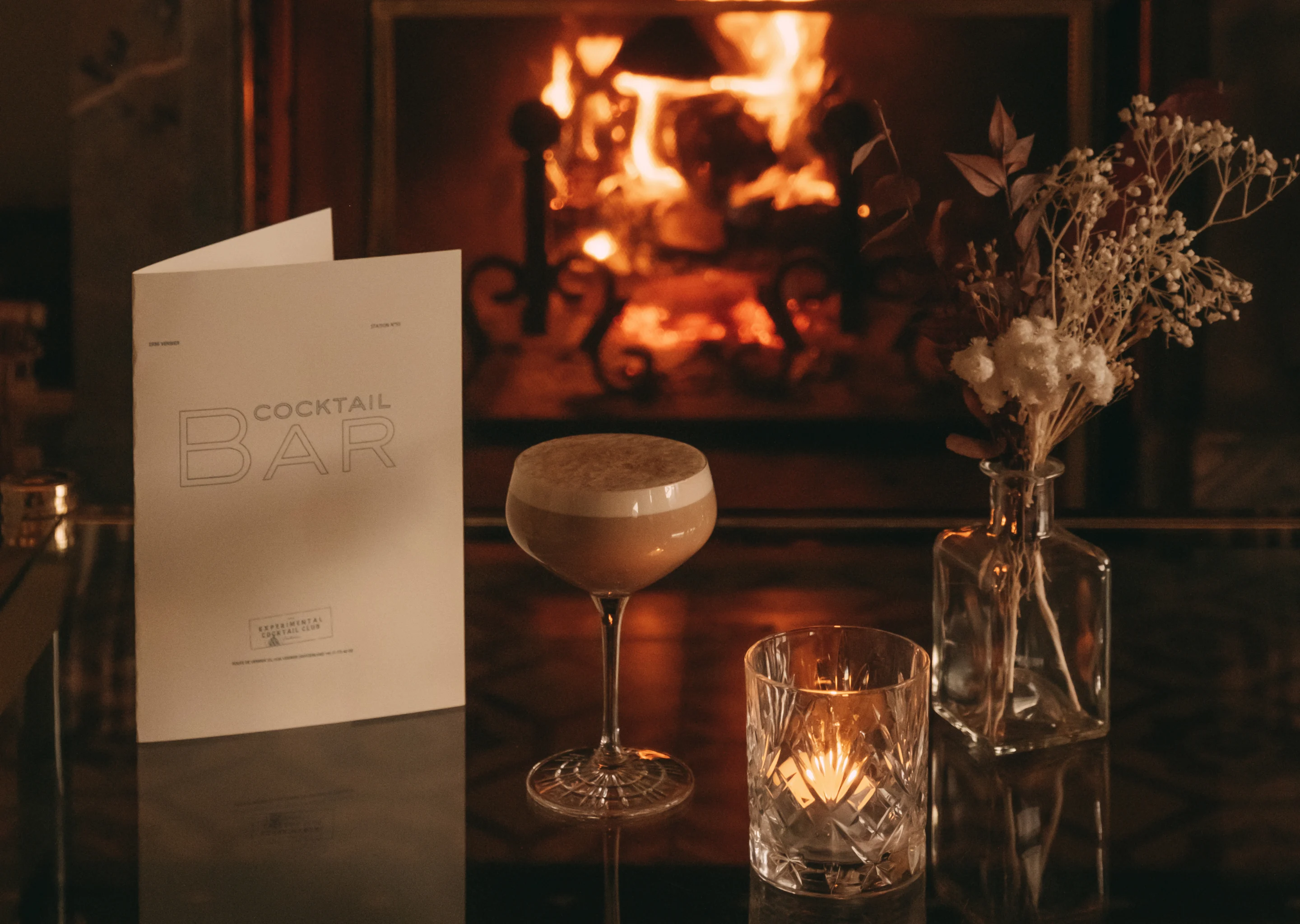 Cocktail glass with frothy drink, lit candle in a glass holder, cocktail bar menu, and dried flower arrangement on a table with a fireplace in the background.