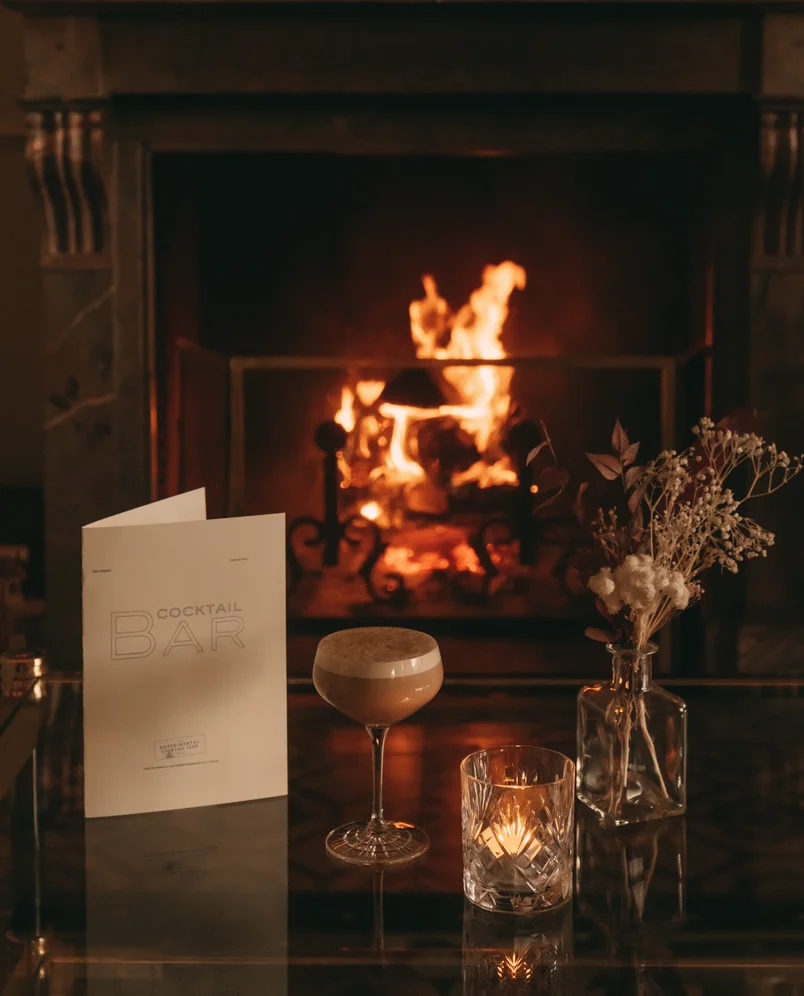Cozy scene with a cocktail glass, lit candle in a crystal holder, dried flowers in a glass vase, and a cocktail bar menu on a table in front of a roaring fireplace.