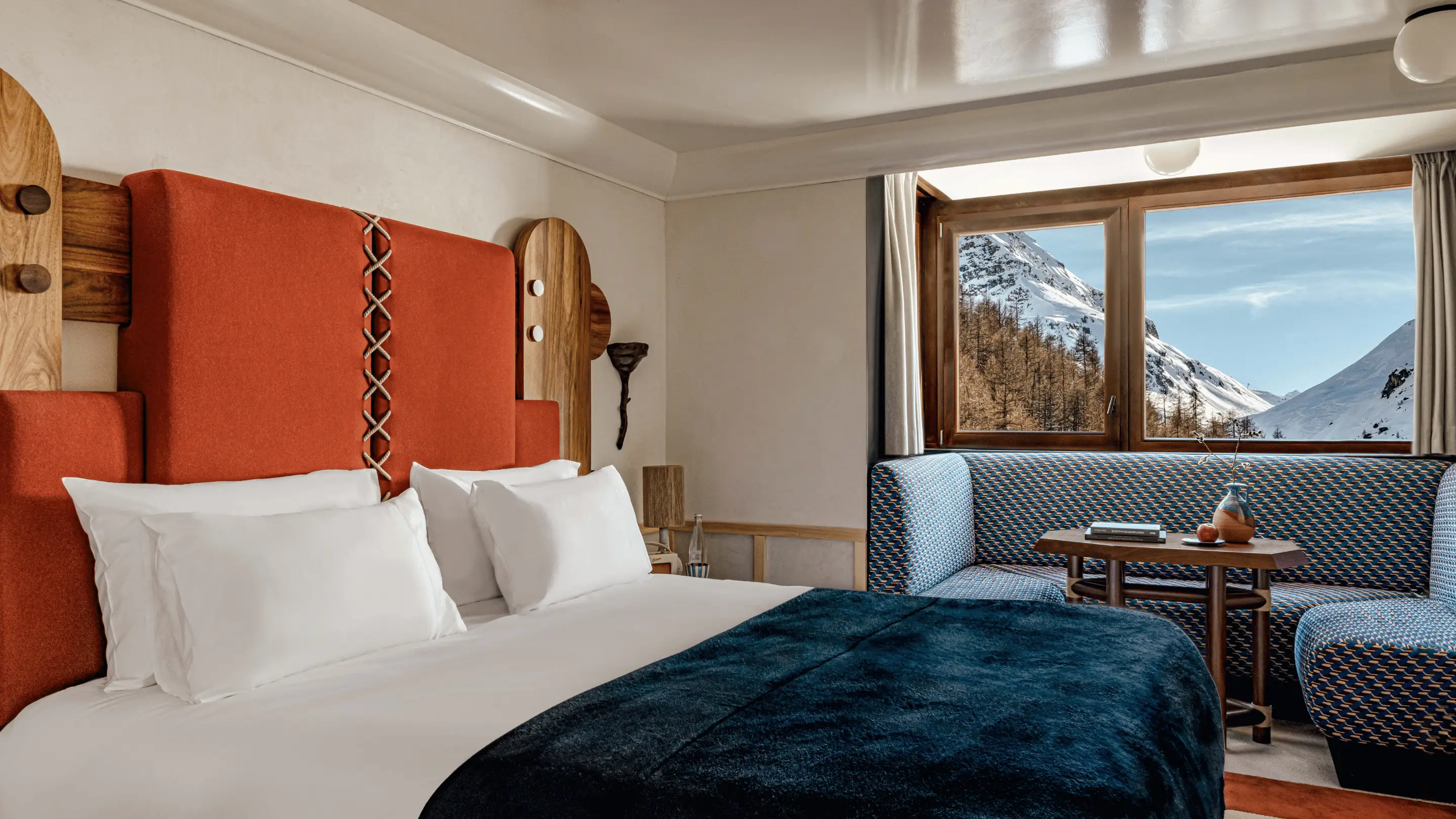 Hotel room with a large bed featuring white pillows and a red-orange headboard, a dark blue blanket, a corner seating area with patterned blue cushions, and a window showing snow-covered mountains.