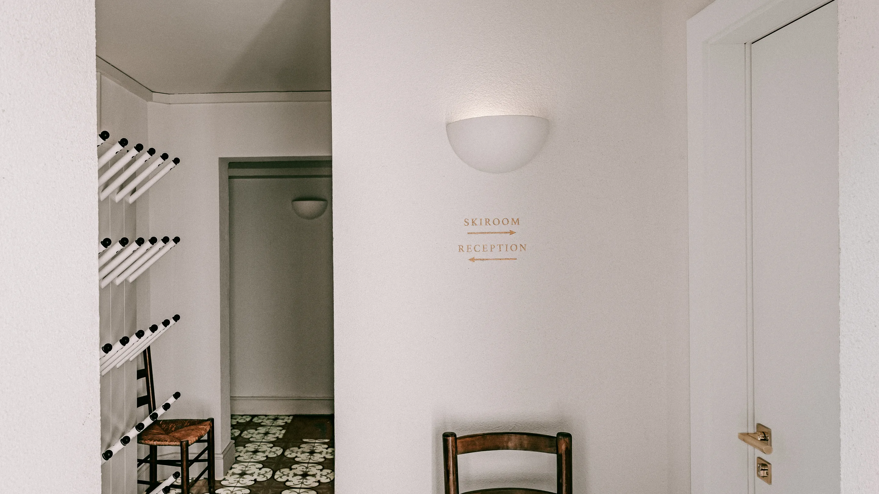 Pasillo minimalista con dos sillas de madera, suelo de baldosas estampadas y letreros en la pared que indican «Sala de esquí» a la derecha y «Recepción» a la izquierda.