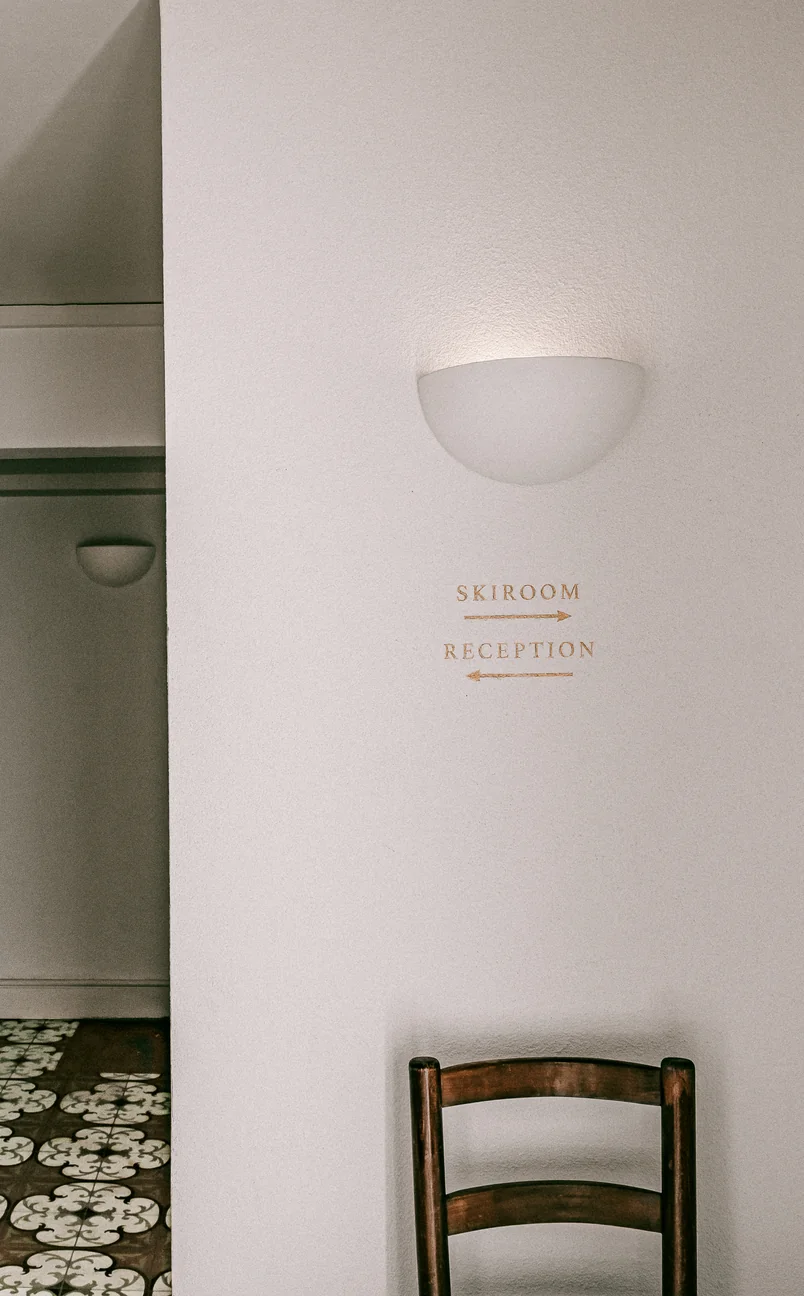 Pared blanca con letrero de color beige que dirige a la sala de esquí a la derecha y a la recepción a la izquierda, encima de una silla de madera y bajo una lámpara de pared.