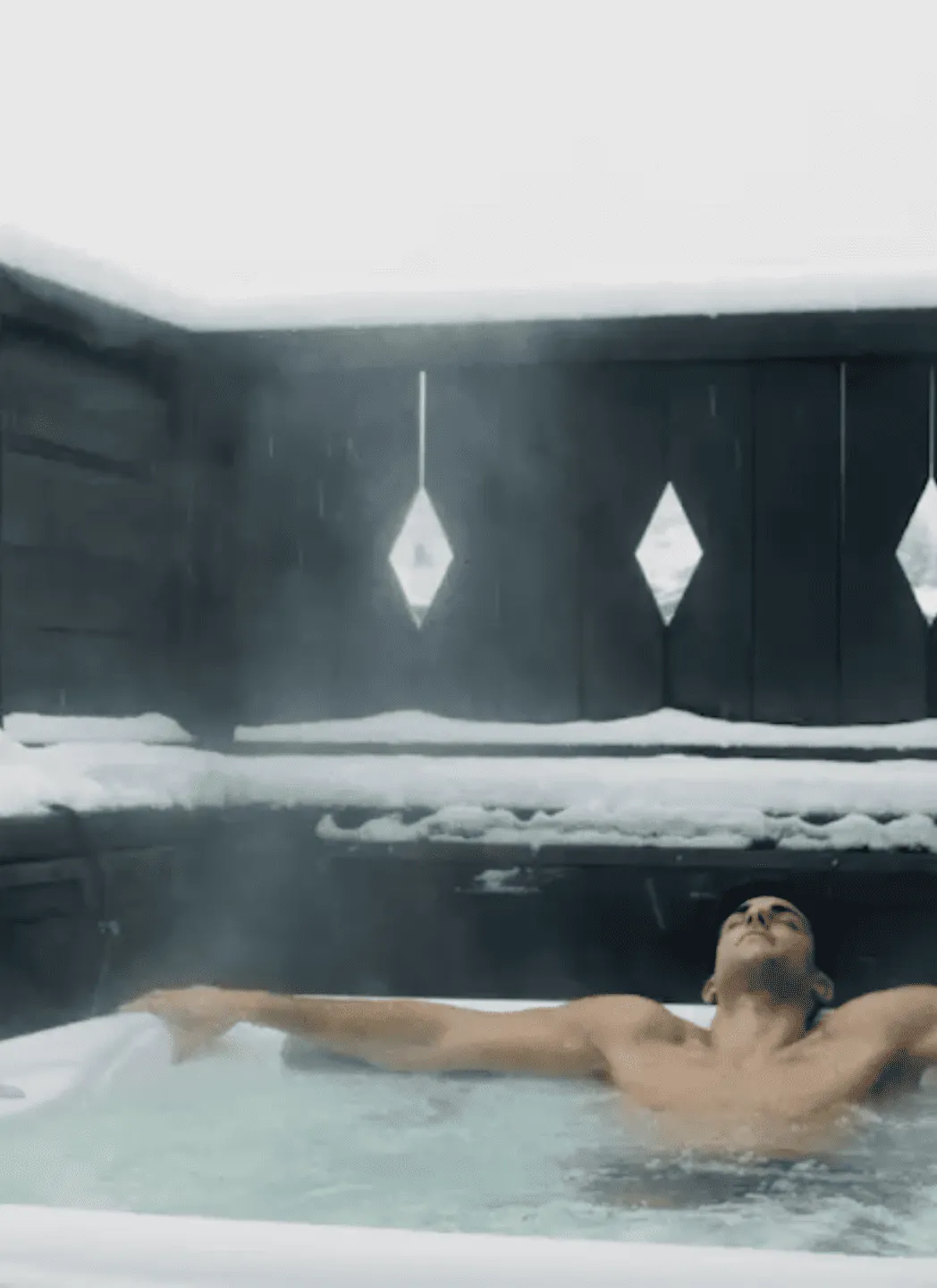 Man relaxing with arms outstretched in an outdoor hot tub surrounded by snow.