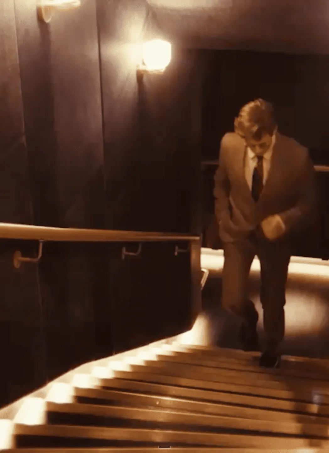 Un hombre de traje sube una escalera con poca luz con luces montadas en la pared y una barandilla.
