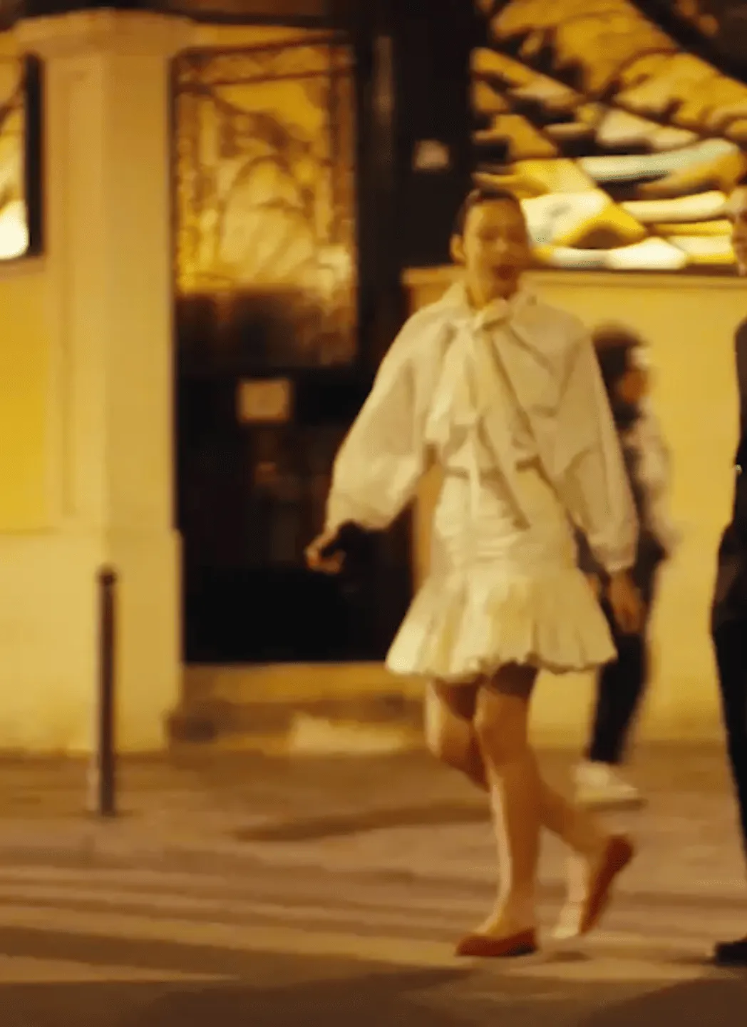 Person wearing a white dress with puffed sleeves and a big bow, walking on a street at night.