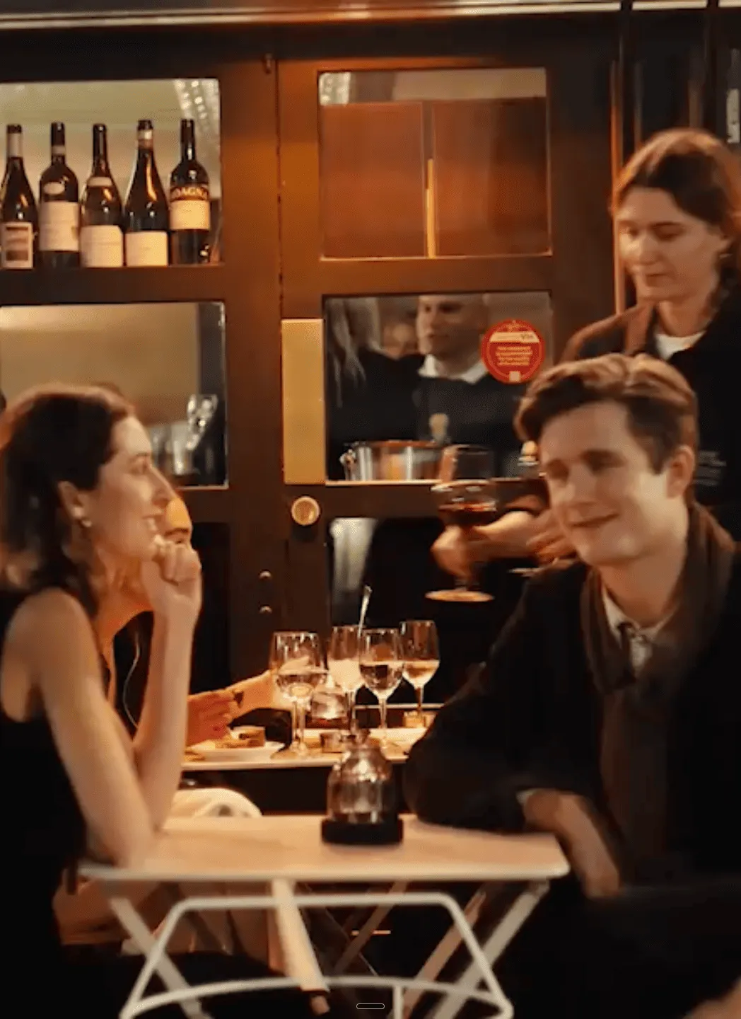 A couple seated at an outdoor café table with wine glasses, being served by a waitress holding a glass of red wine.