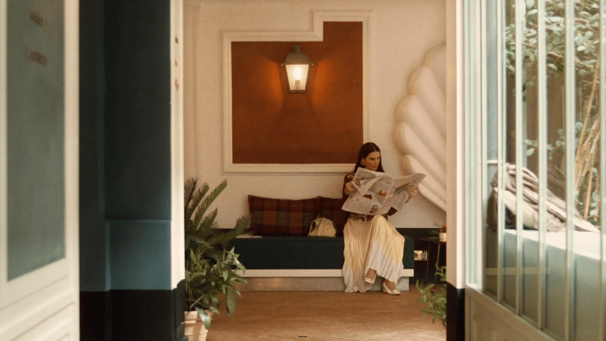 Woman sitting on a bench reading a newspaper under a wall lantern in a cozy indoor space with plants.