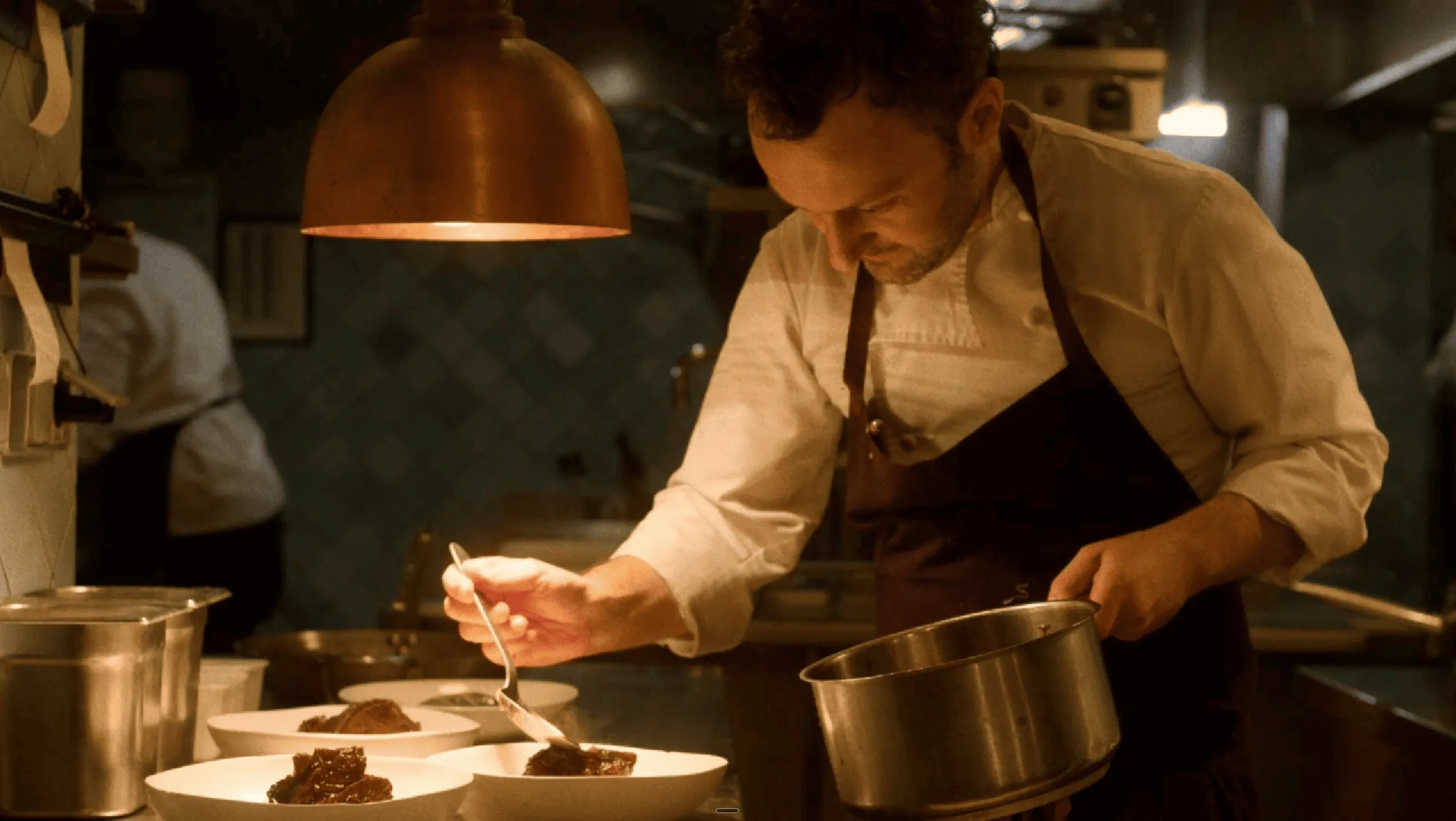 Chef in white shirt and dark apron carefully plating food in a dimly lit kitchen under a copper light.