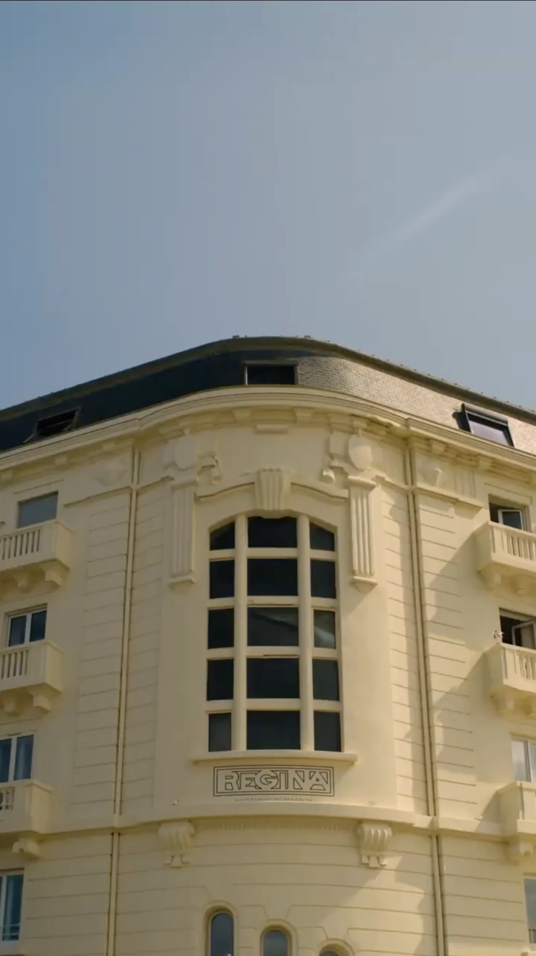 Upper corner of a beige historic building with decorative columns, balconies, a large central window, and the name 'REGINA' below the window under a clear blue sky.