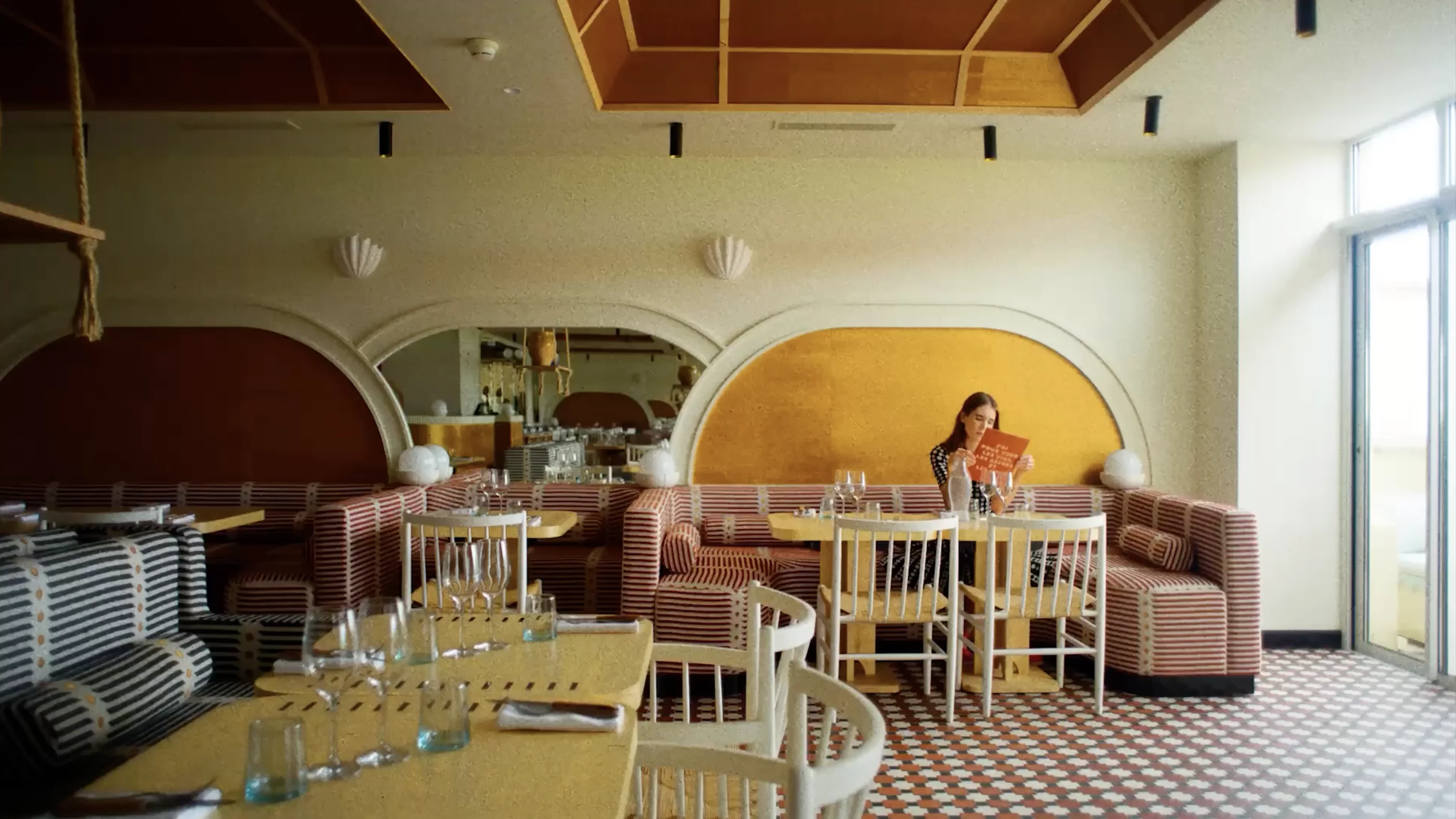 Woman sitting alone on a striped bench in a stylish restaurant, looking at a menu with sunlight coming through large windows.