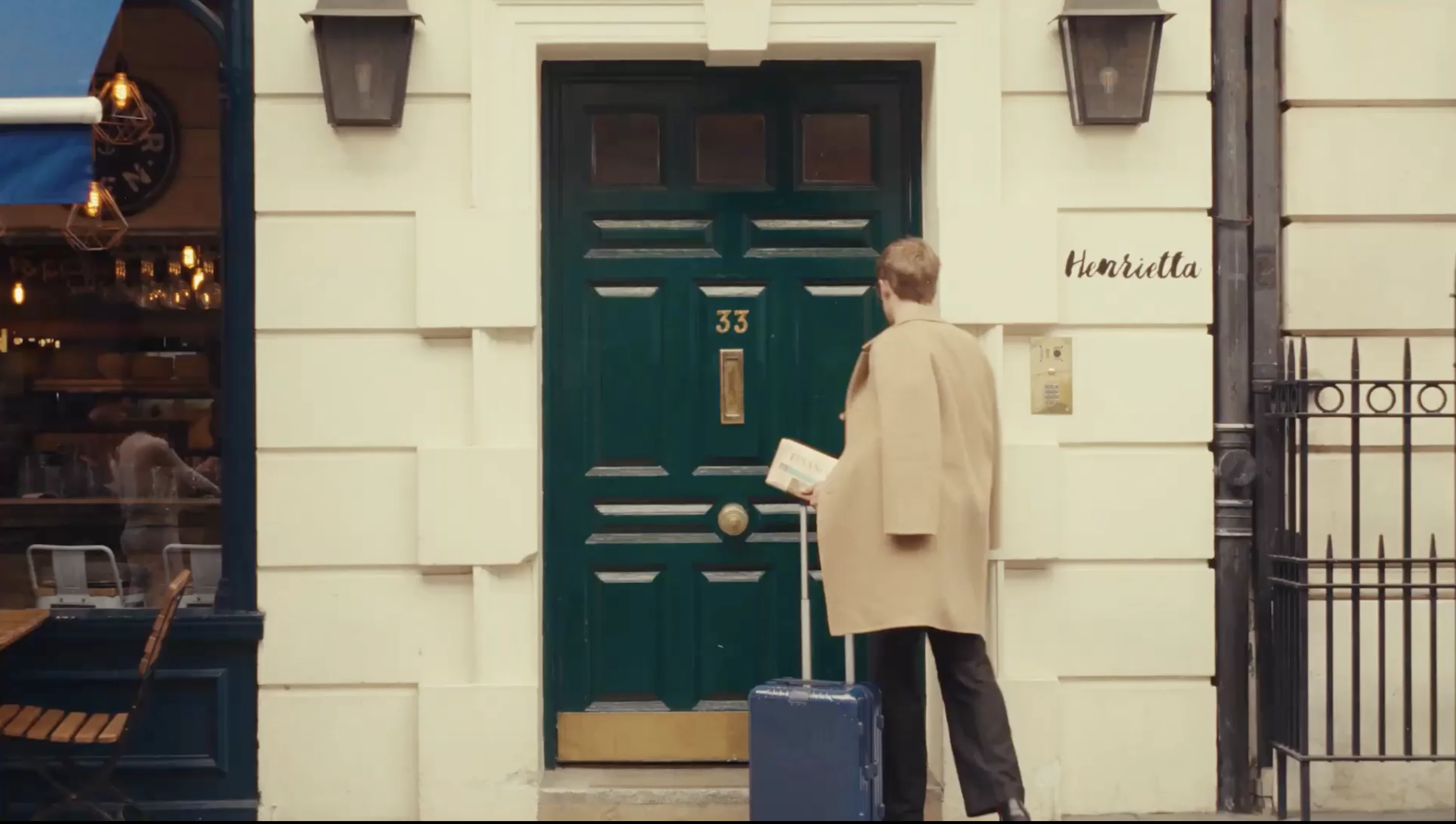 Person in beige coat holding a book and a suitcase standing in front of a dark green door numbered 33 with the name Henrietta nearby.
