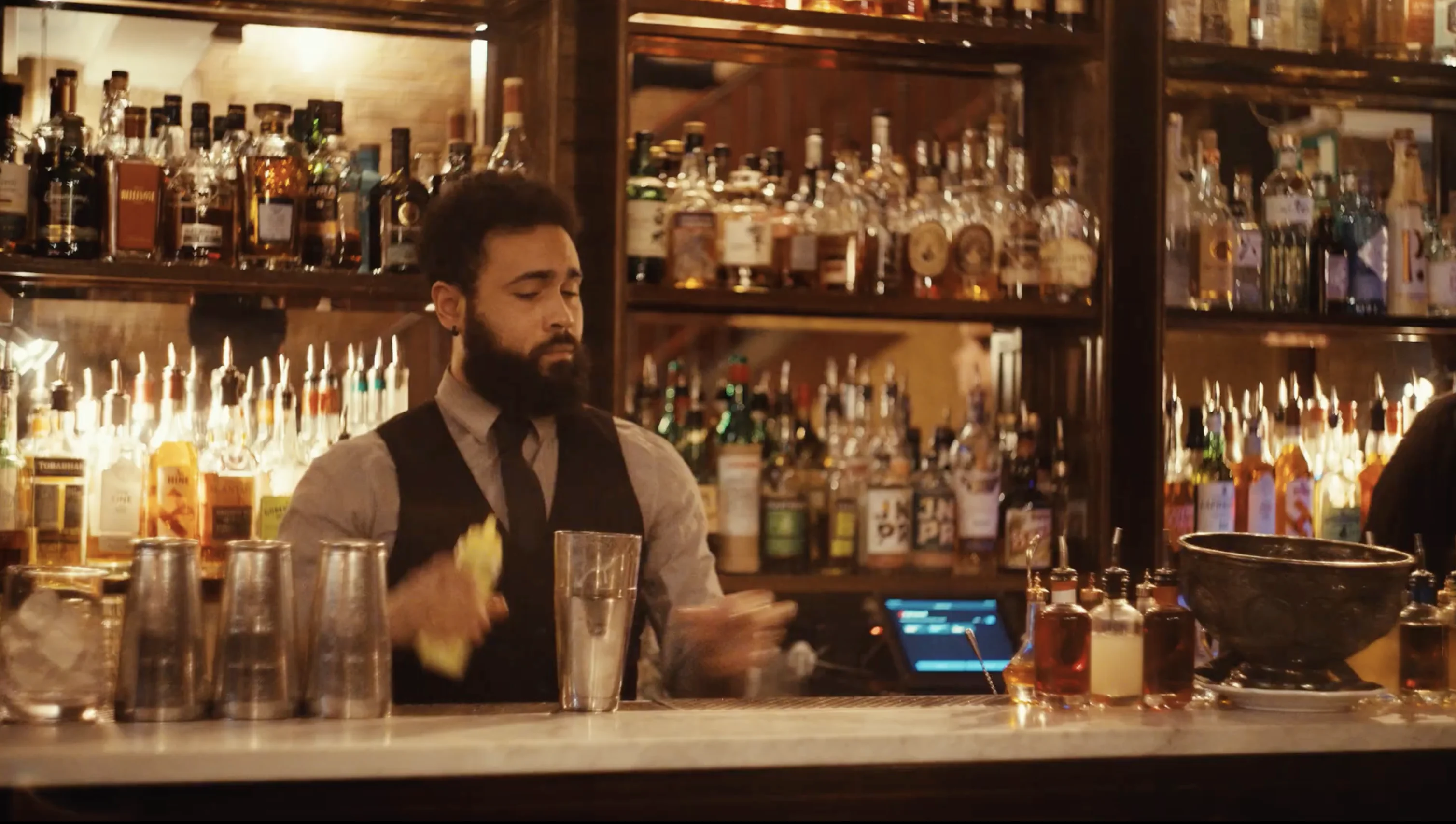 Bartender with a beard shaking a cocktail shaker behind a bar stocked with various liquor bottles.