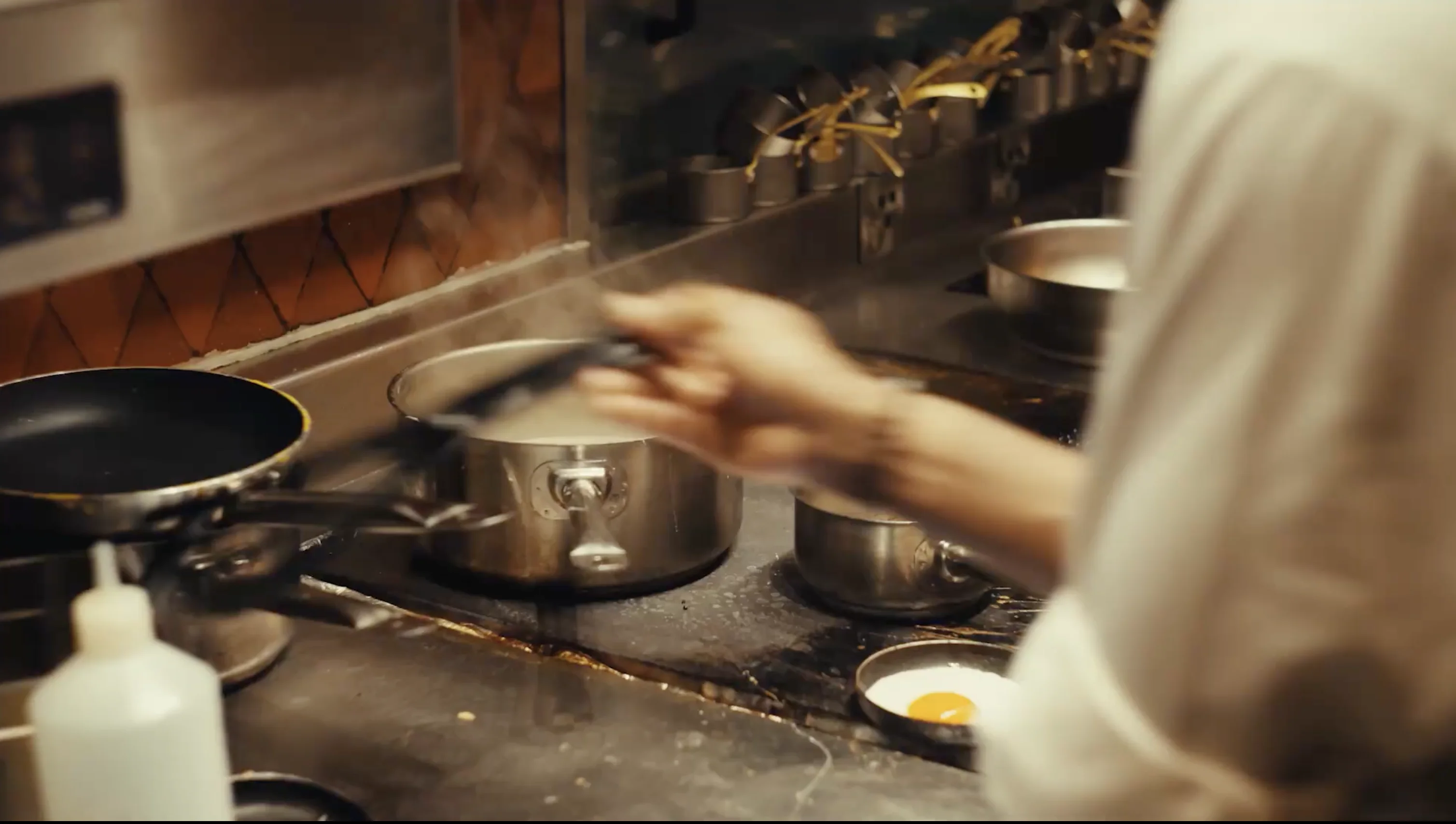 Person cooking at a stove with pans and pots, one pan containing a fried egg yolk.