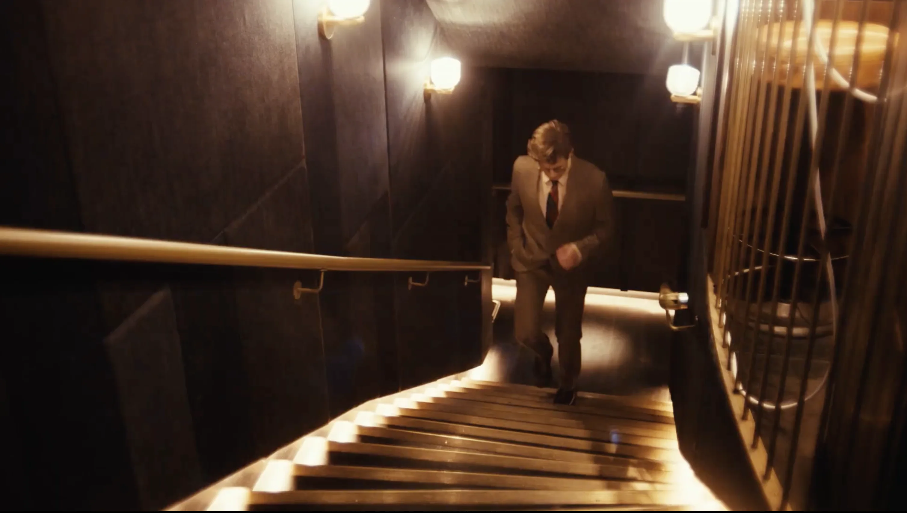 Man in a suit walking up a dimly lit staircase with warm wall lights and a brass handrail.