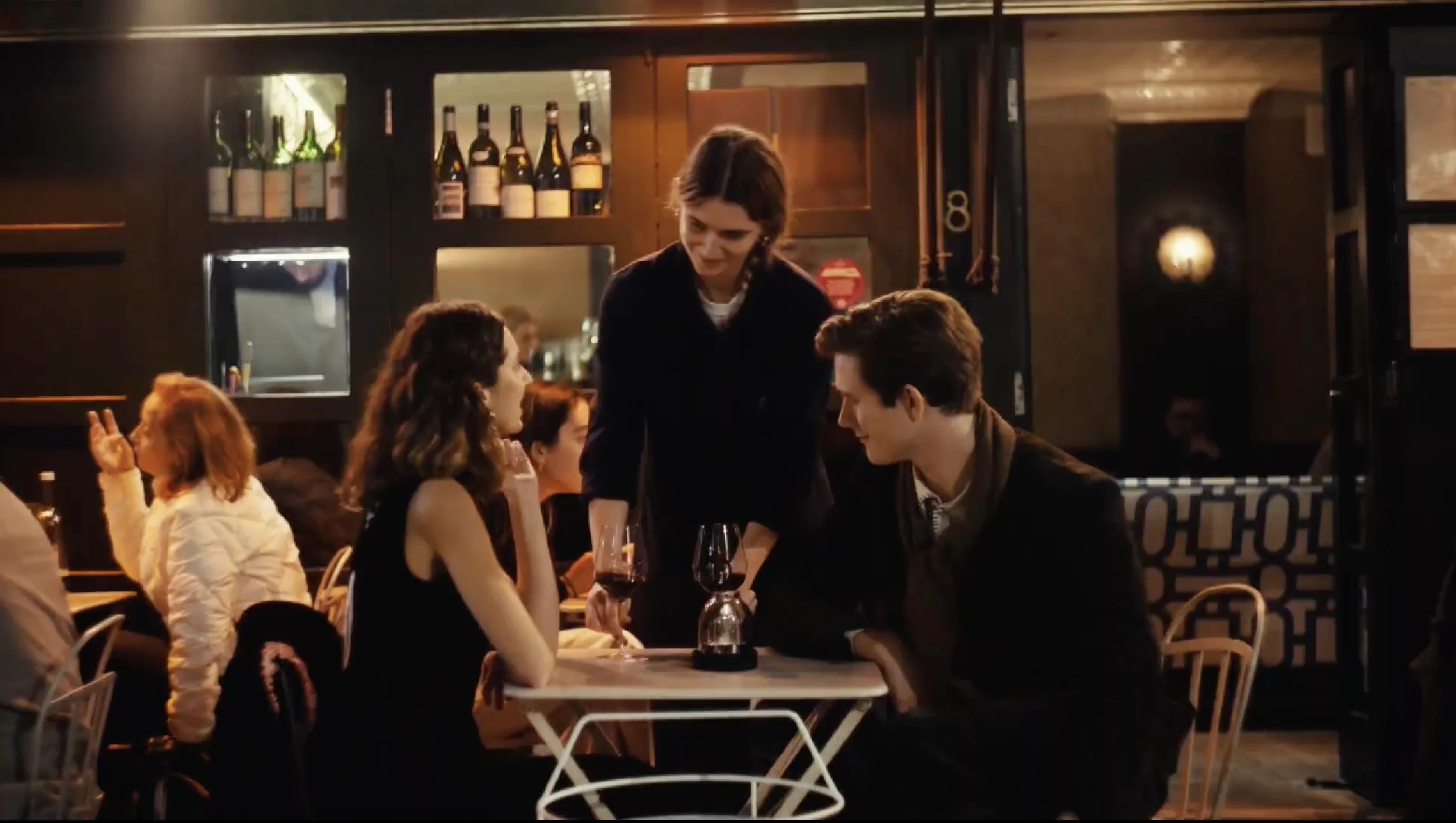 A waiter serving wine to a couple seated at a small table in a cozy restaurant with bottles displayed behind the bar.