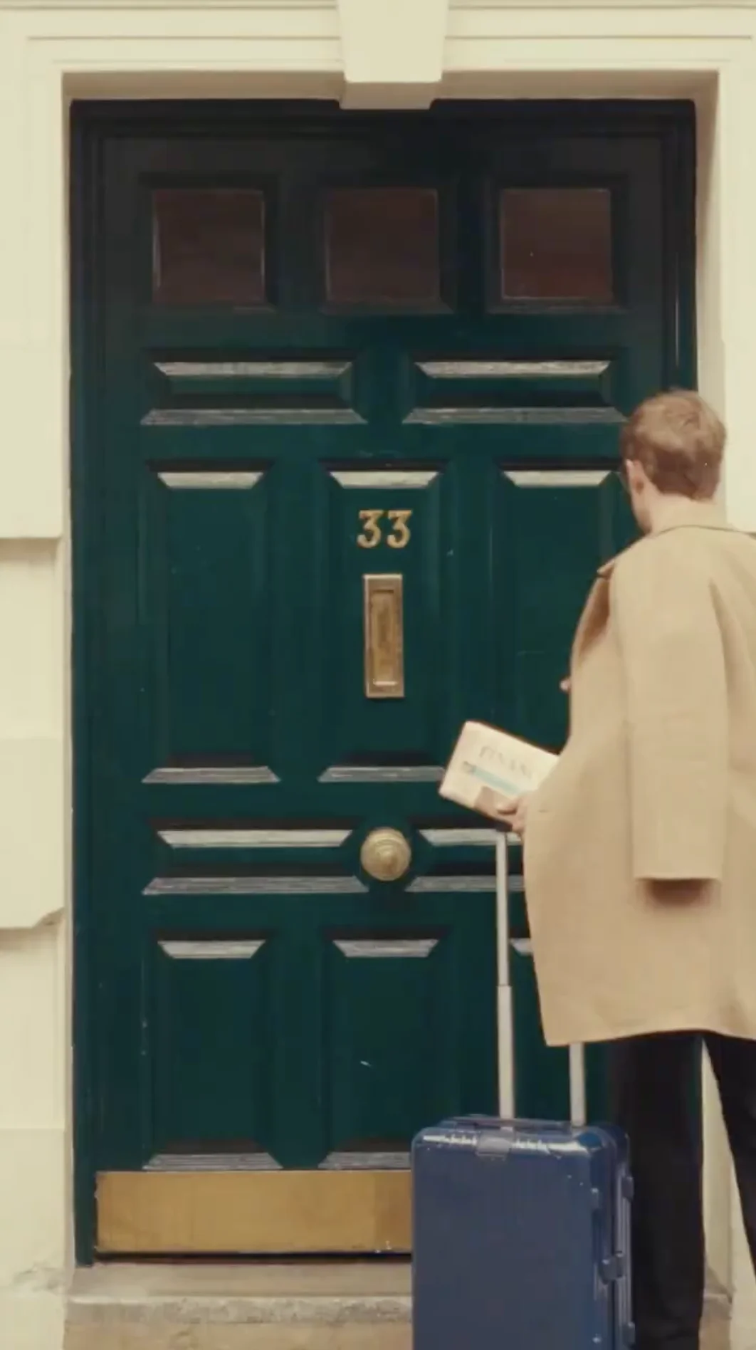 Person in beige coat holding a newspaper and rolling a blue suitcase in front of a dark green door numbered 33.