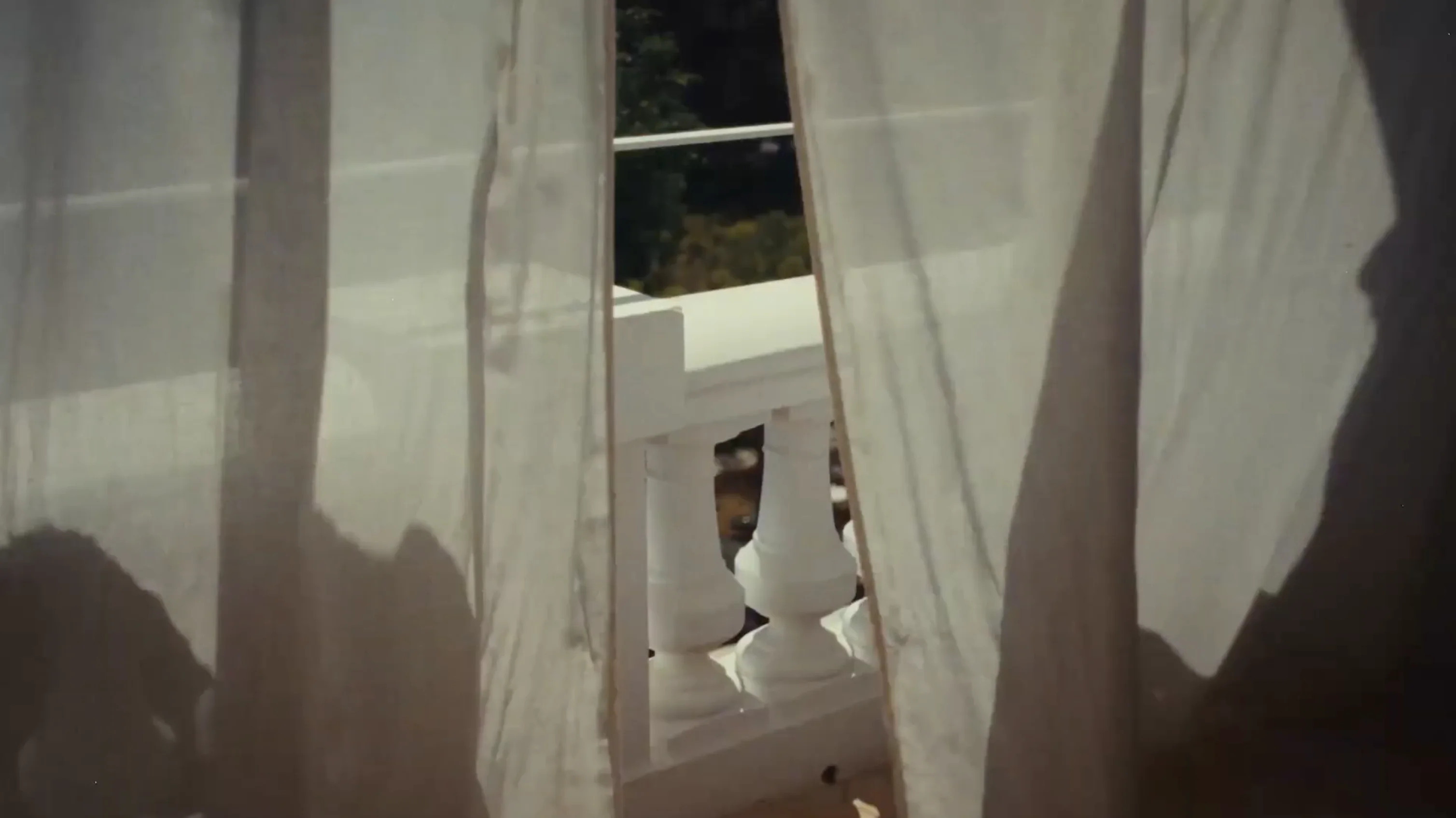 View through sheer white curtains showing white balcony balusters and greenery outside.