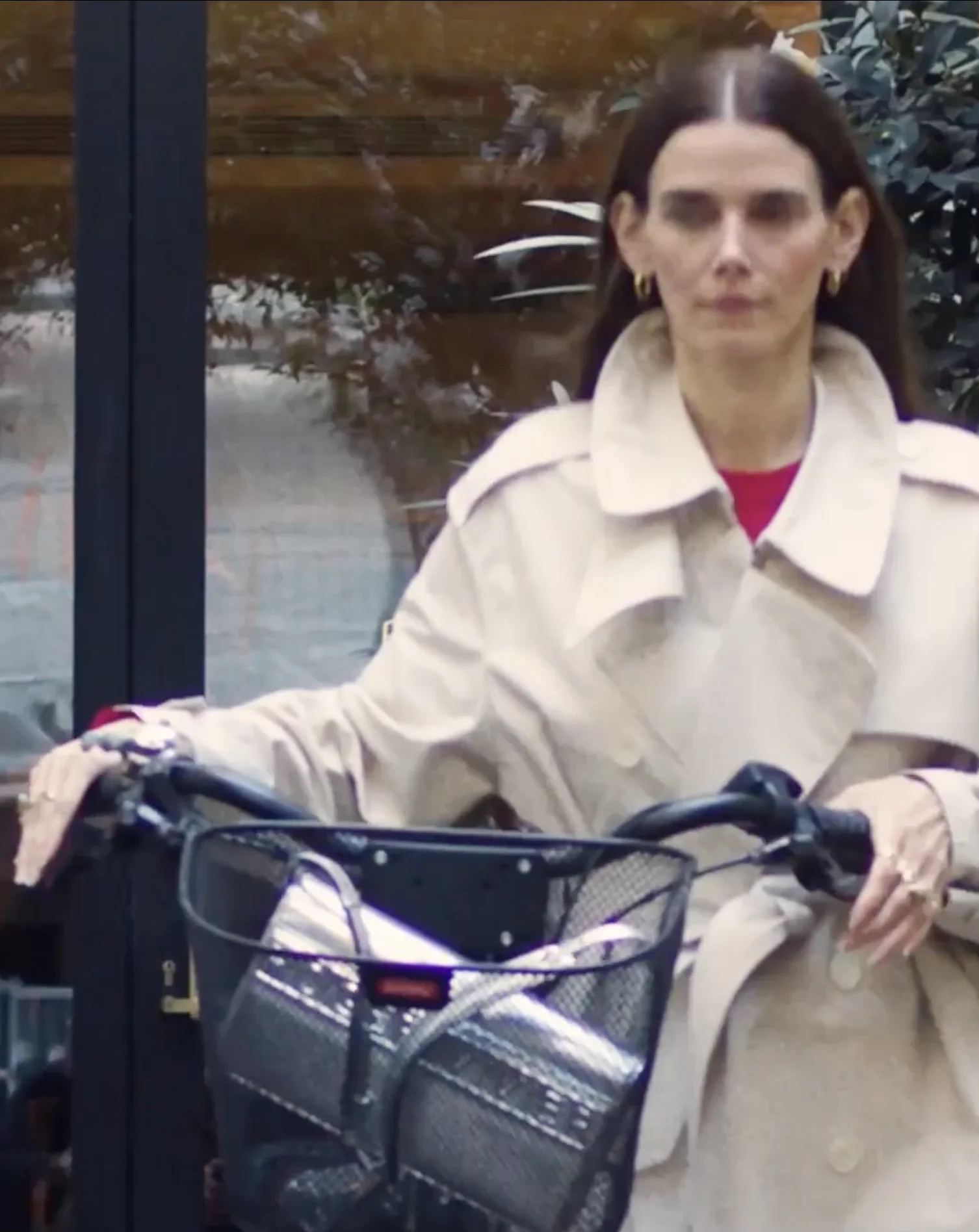 Woman in beige trench coat holding the handlebars of a bicycle with a front basket.