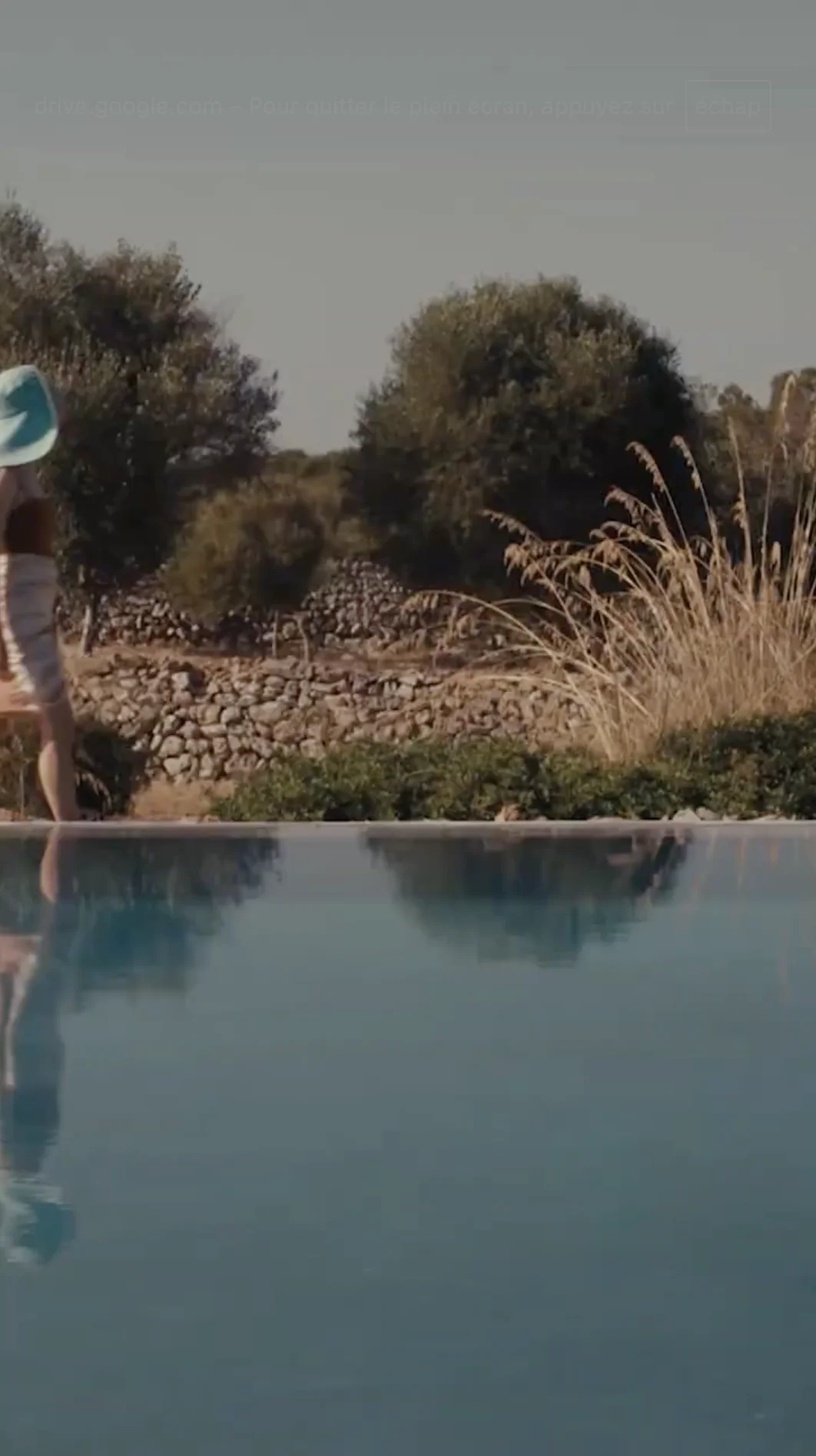 Person in a white dress and blue sunhat standing near a pool with stone walls and trees in the background.