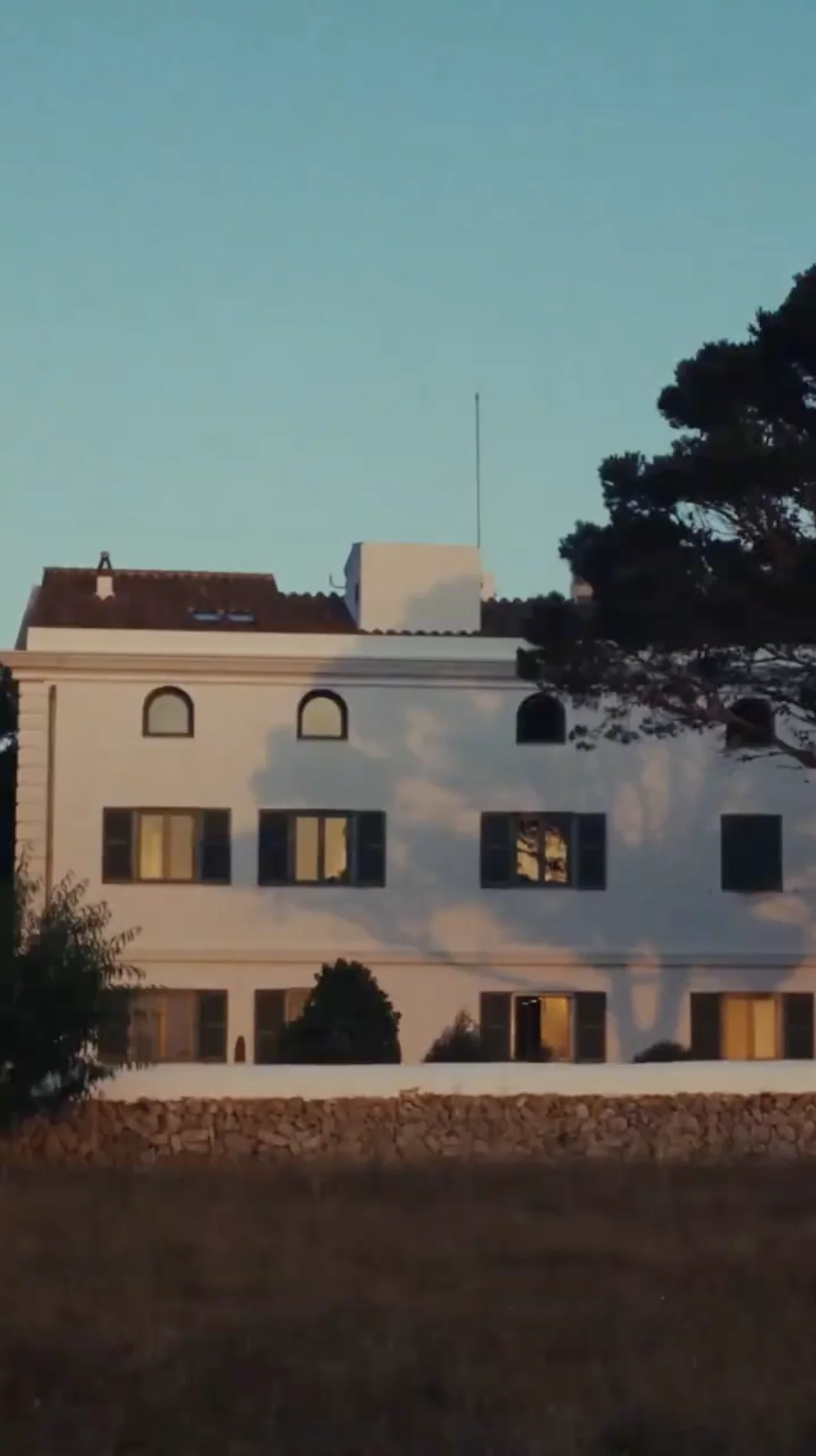 White multi-story house with arched windows and green shutters, casting tree shadows at sunset.