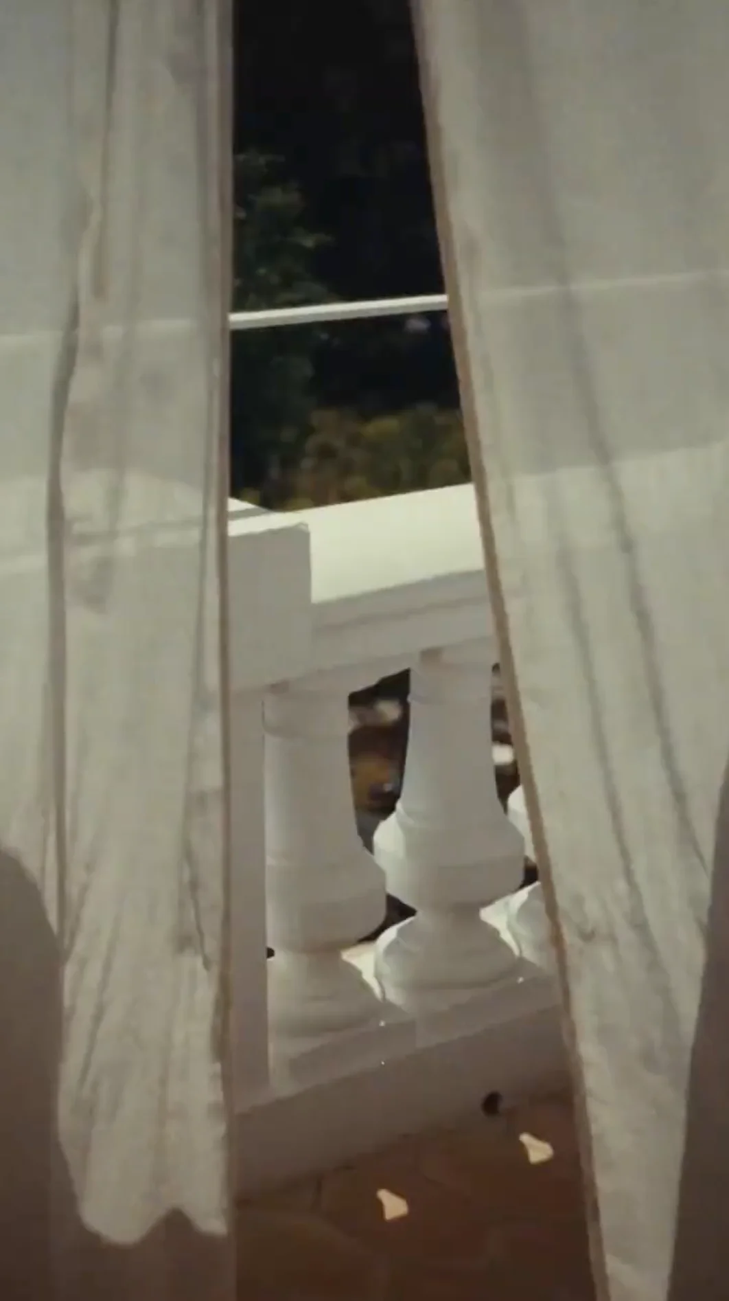 View through light curtains of white balusters on a balcony with dark greenery in the background.