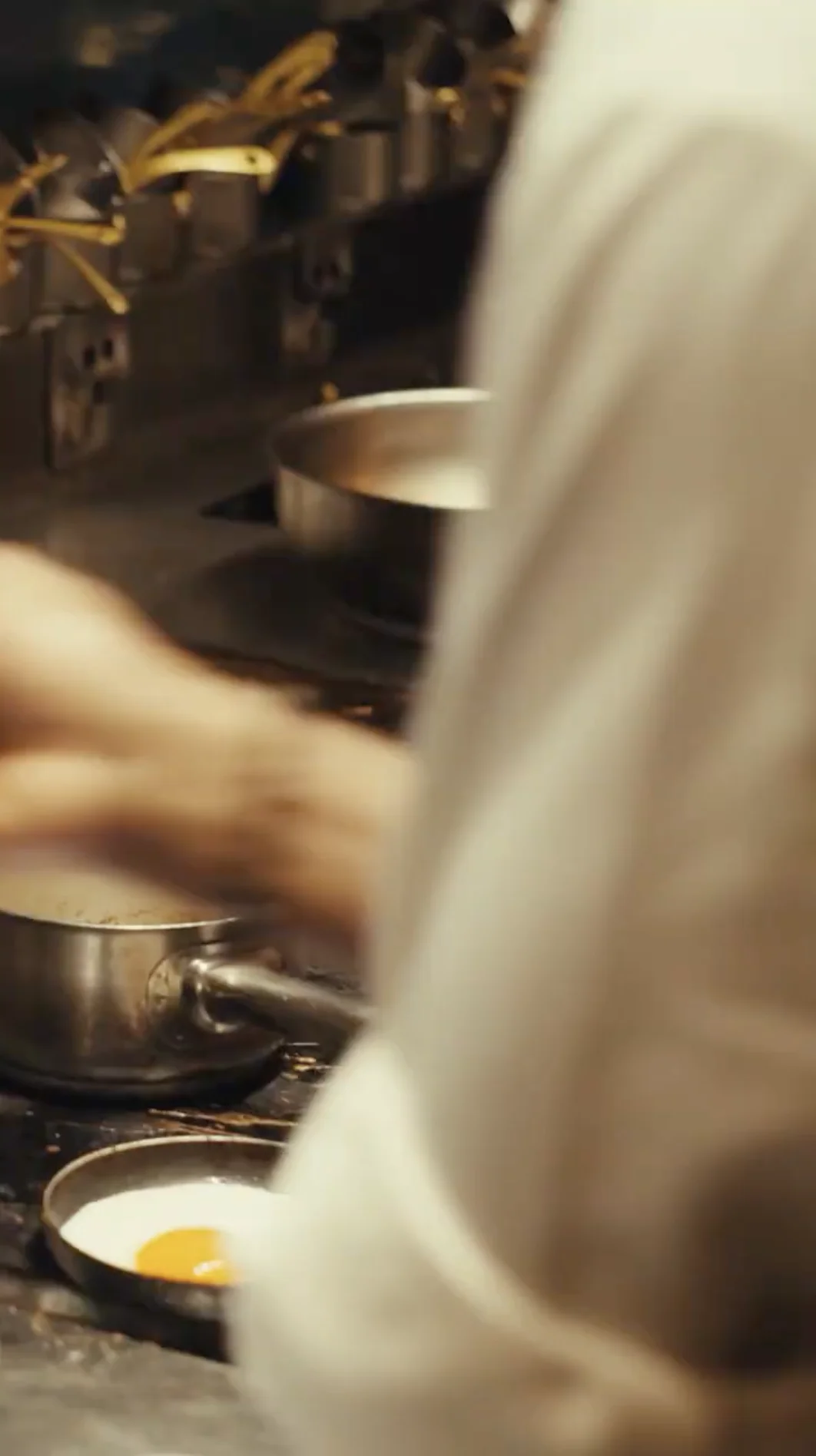 Person cooking an egg in a frying pan on a stovetop with other pans in the background.