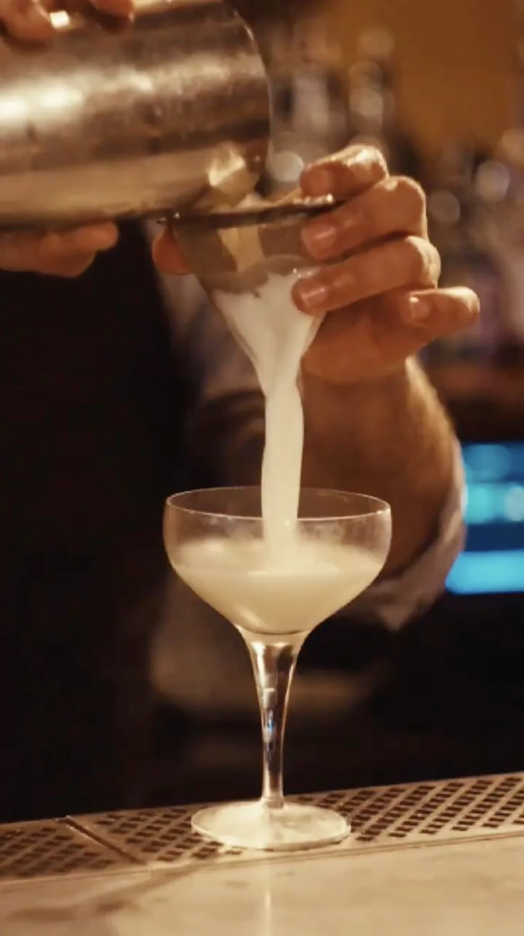 Bartender pouring a frothy white cocktail through a strainer into a coupe glass.