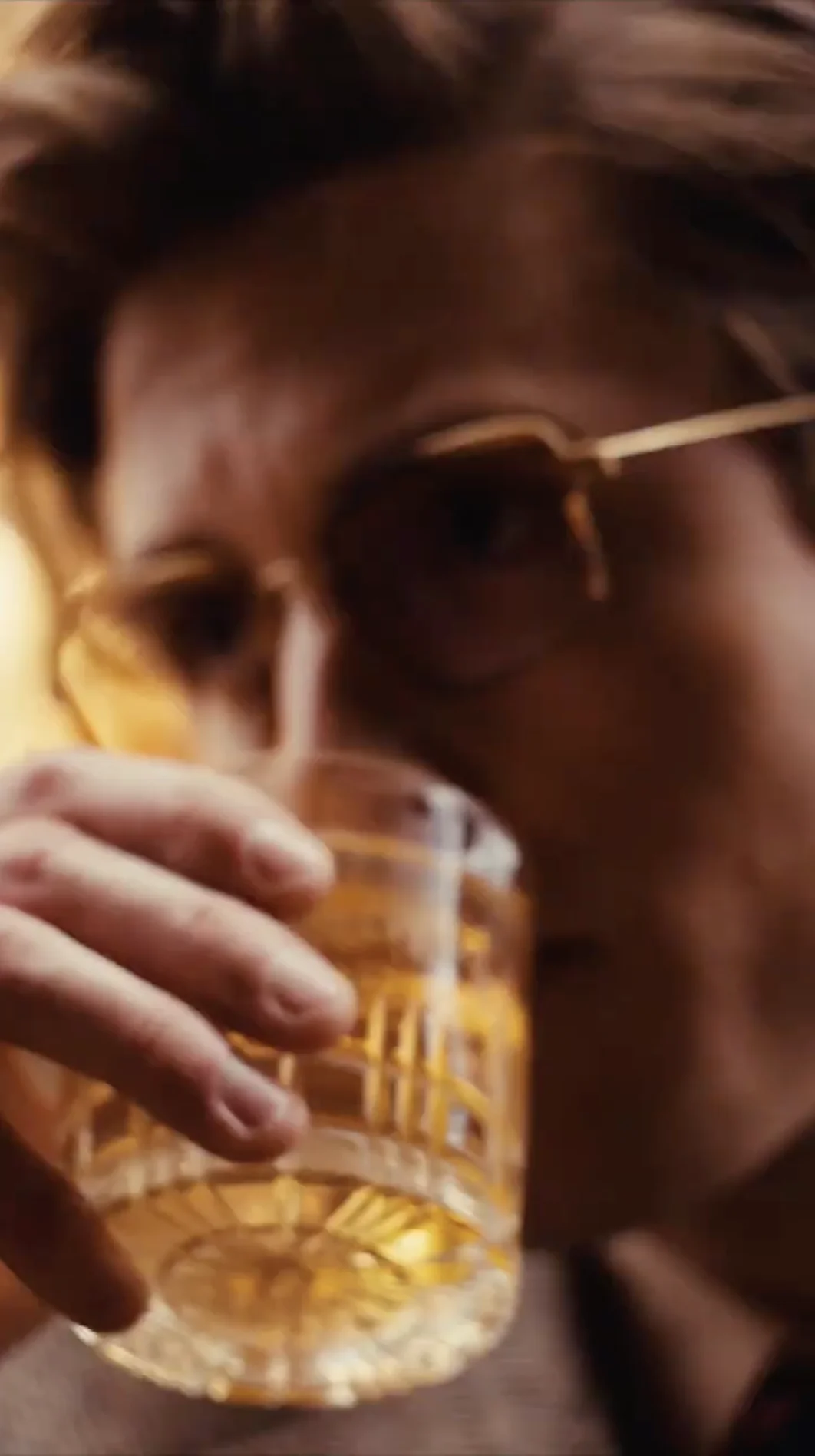 Person wearing glasses holding and drinking from a textured glass with an amber-colored liquid.