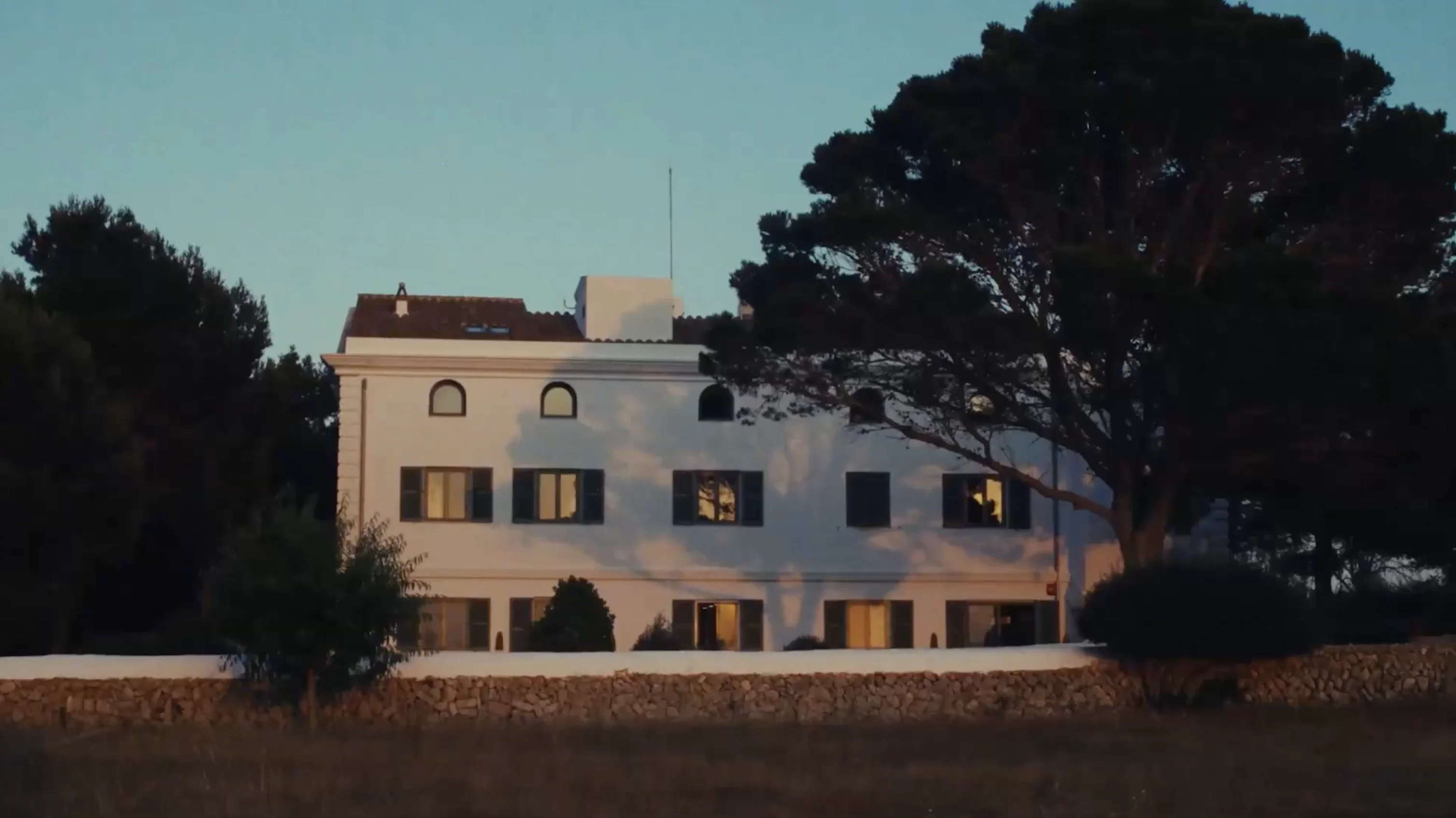 White multi-story house with dark shutters partially shaded by large trees during sunset.