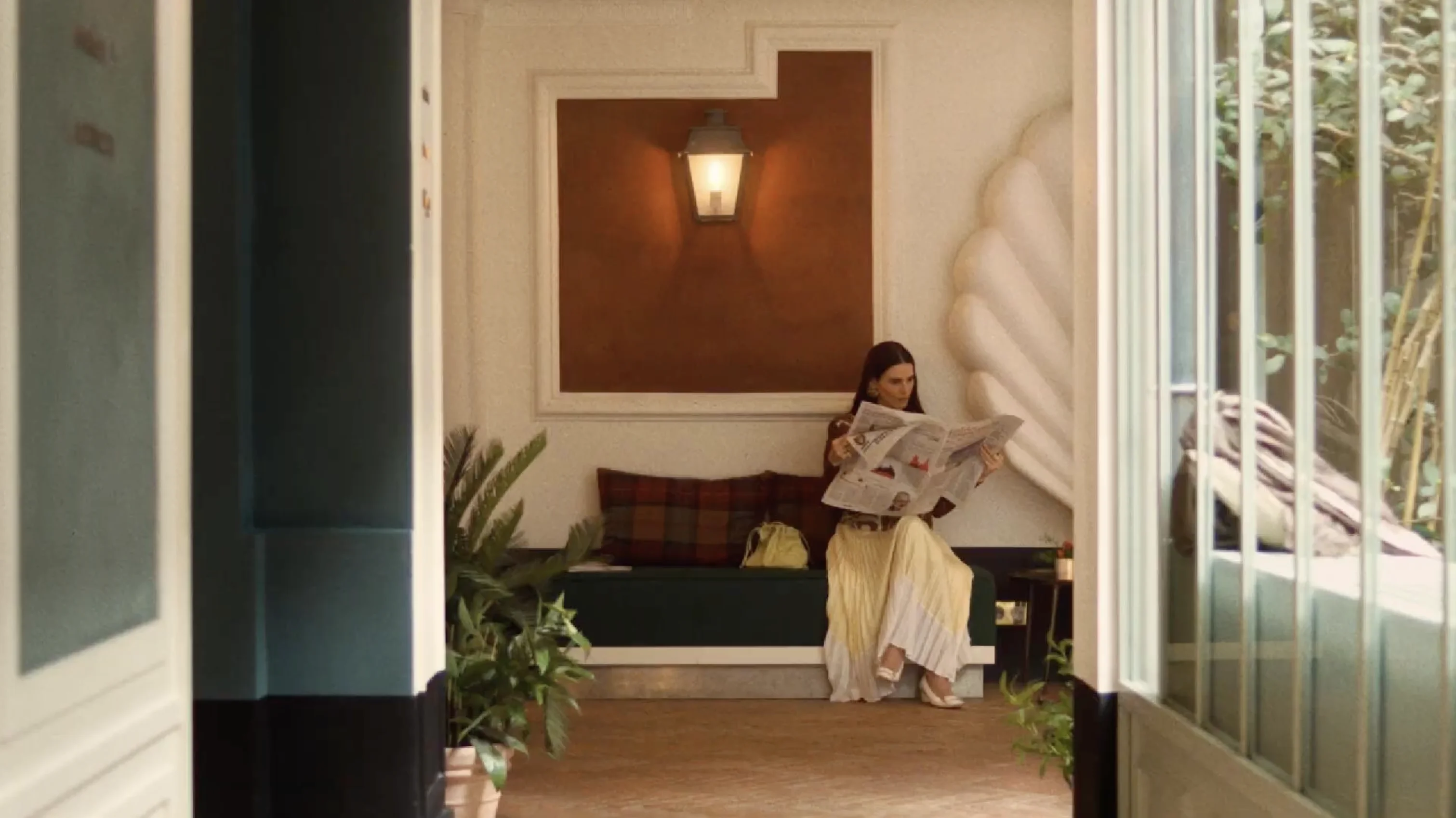 Woman sitting on a bench indoors reading a newspaper with plants and a wall lamp in the background.