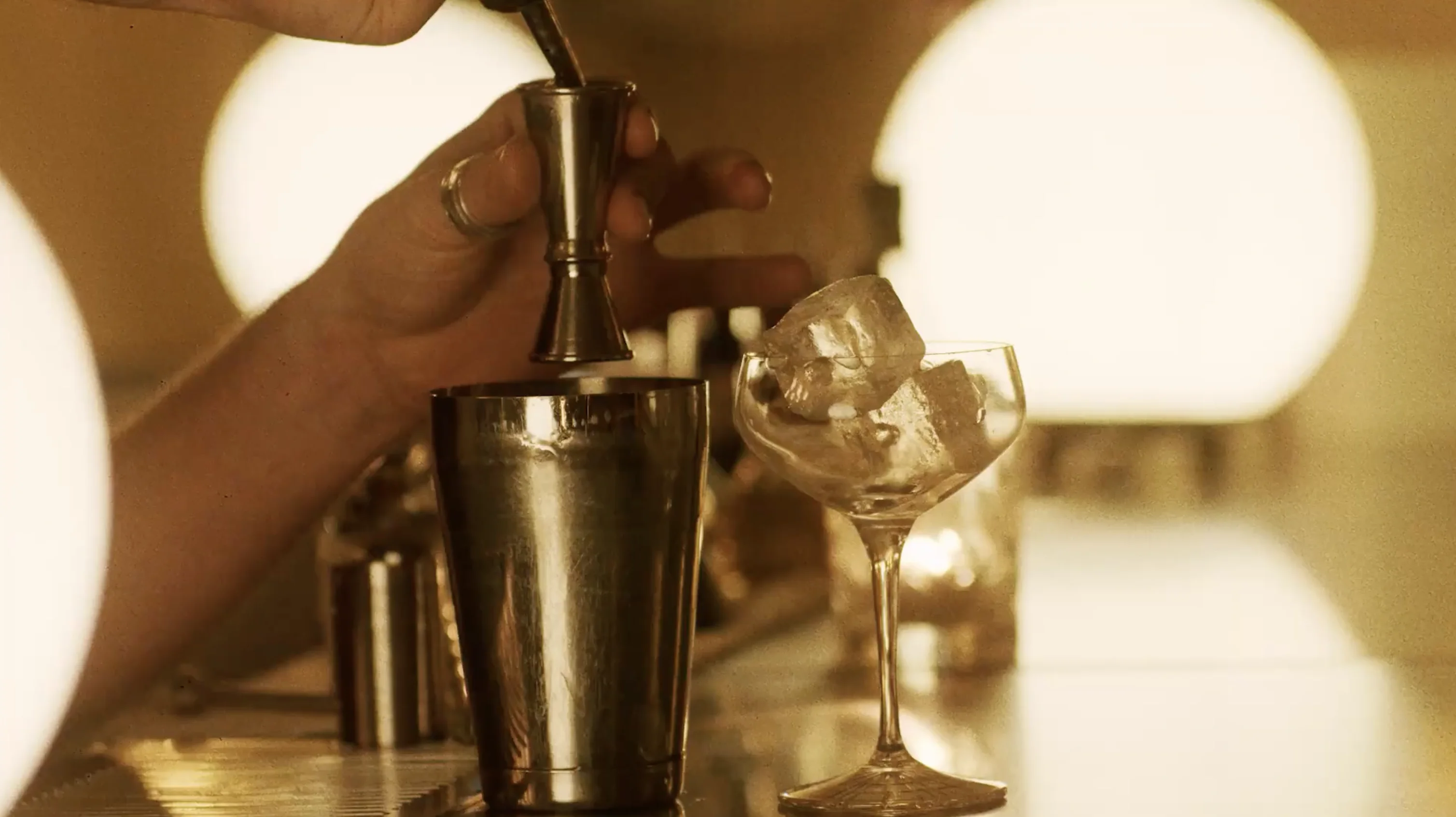 Hand holding a jigger pouring liquid into a metal cocktail shaker next to a glass with ice cubes on a bar counter.