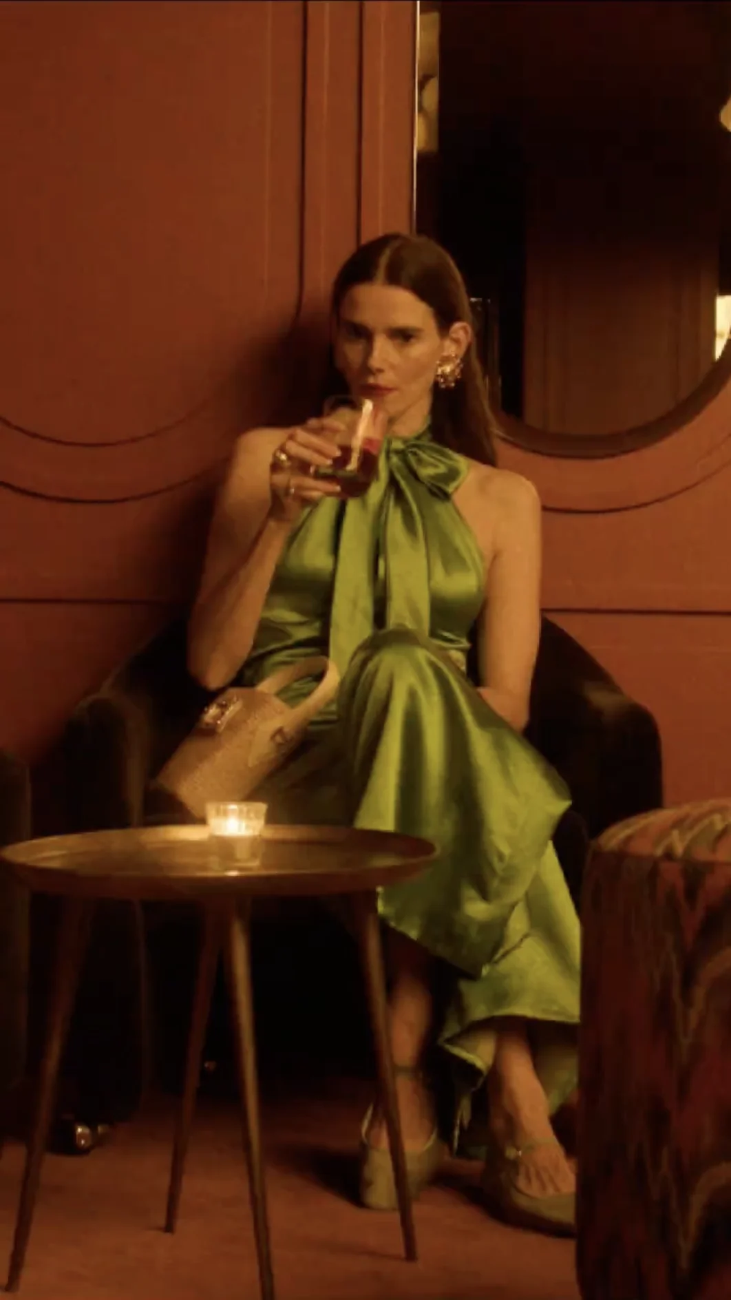 Woman in a green satin dress seated in a chair holding a glass with a drink, next to a small round table with a lit candle.