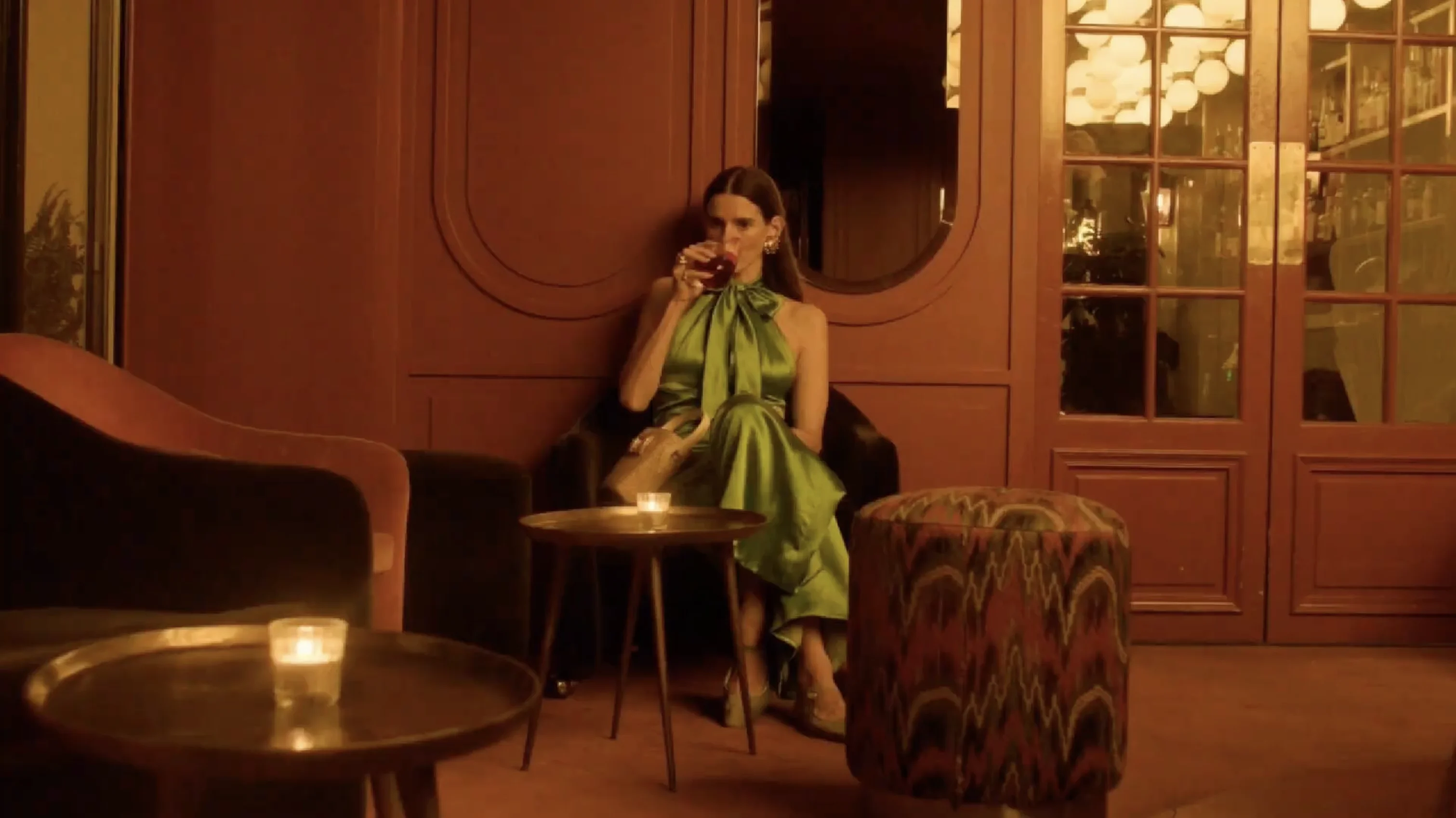 Woman in a green satin dress sitting in a dimly lit lounge drinking a cocktail.